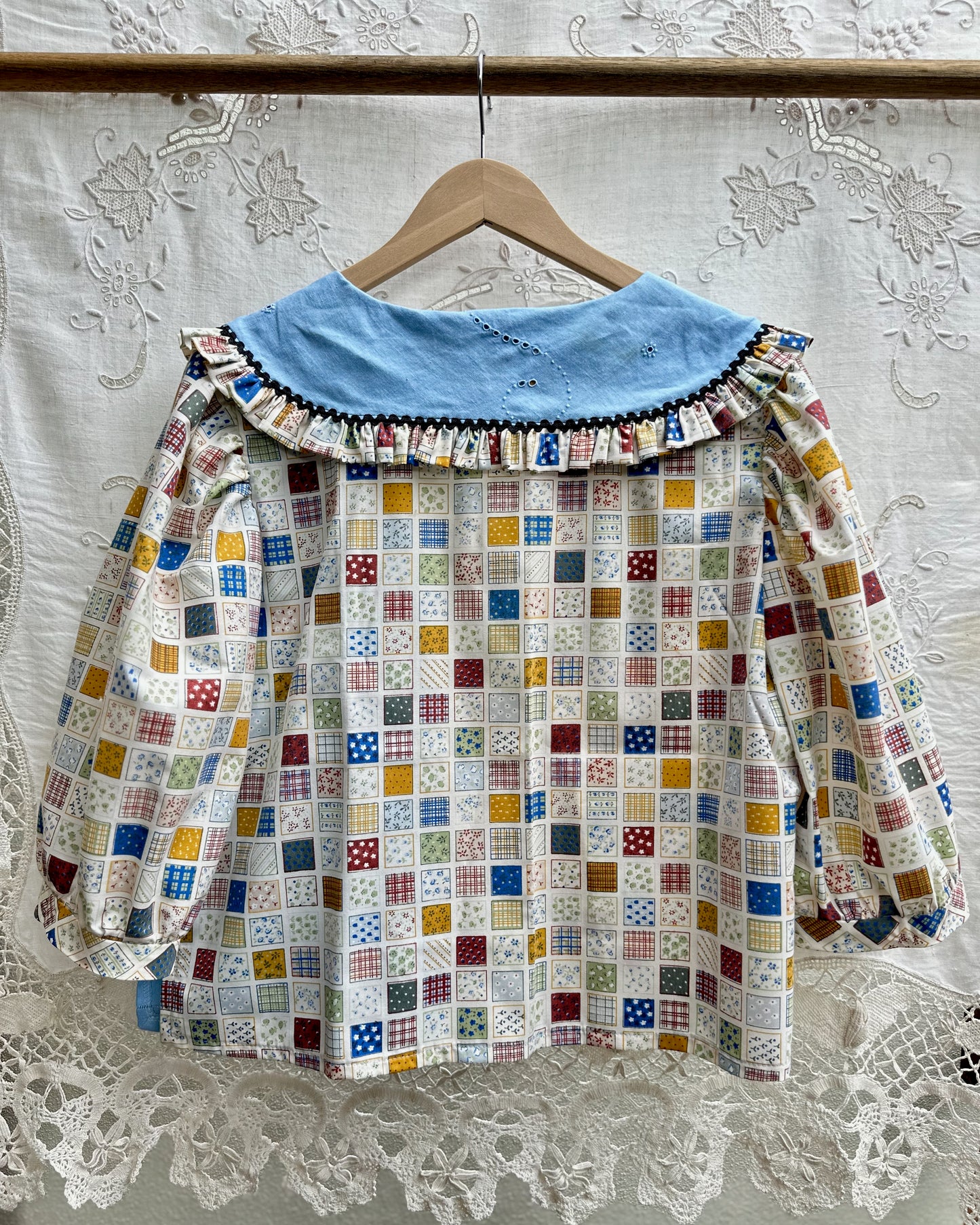 A vintage-style long-sleeve blouse hanging on a wooden hanger against a white lace curtain backdrop. The blouse features a colorful patchwork grid pattern on the body and a solid light blue collar with a ruffled trim.