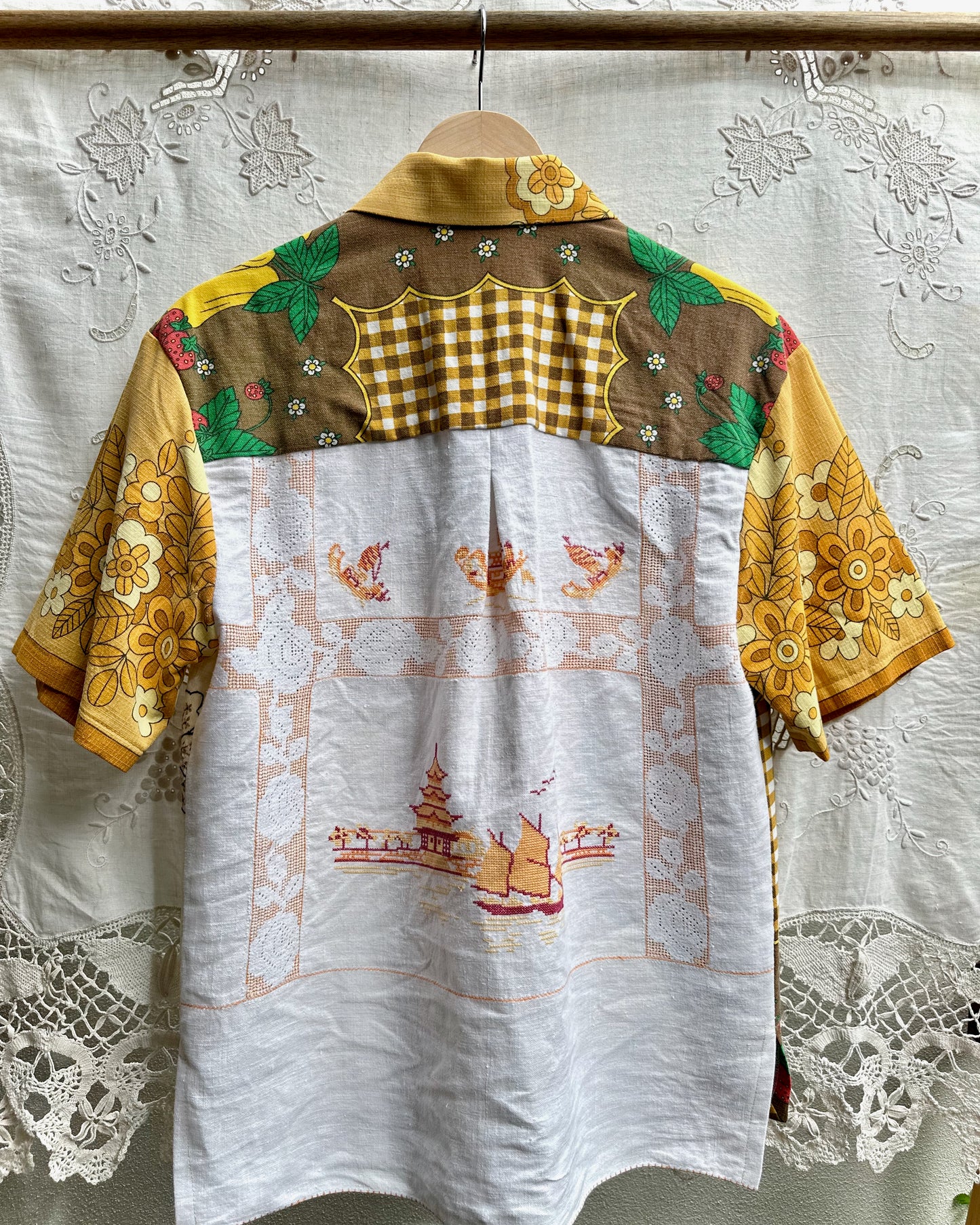 A short-sleeve button-up shirt hanging on a wooden hanger against a white lace curtain. The shirt features a complex multicolor design: a white lower half with orange temple embroidery, a brown and yellow gingham yoke, and yellow floral sleeves with green leaf accents.
