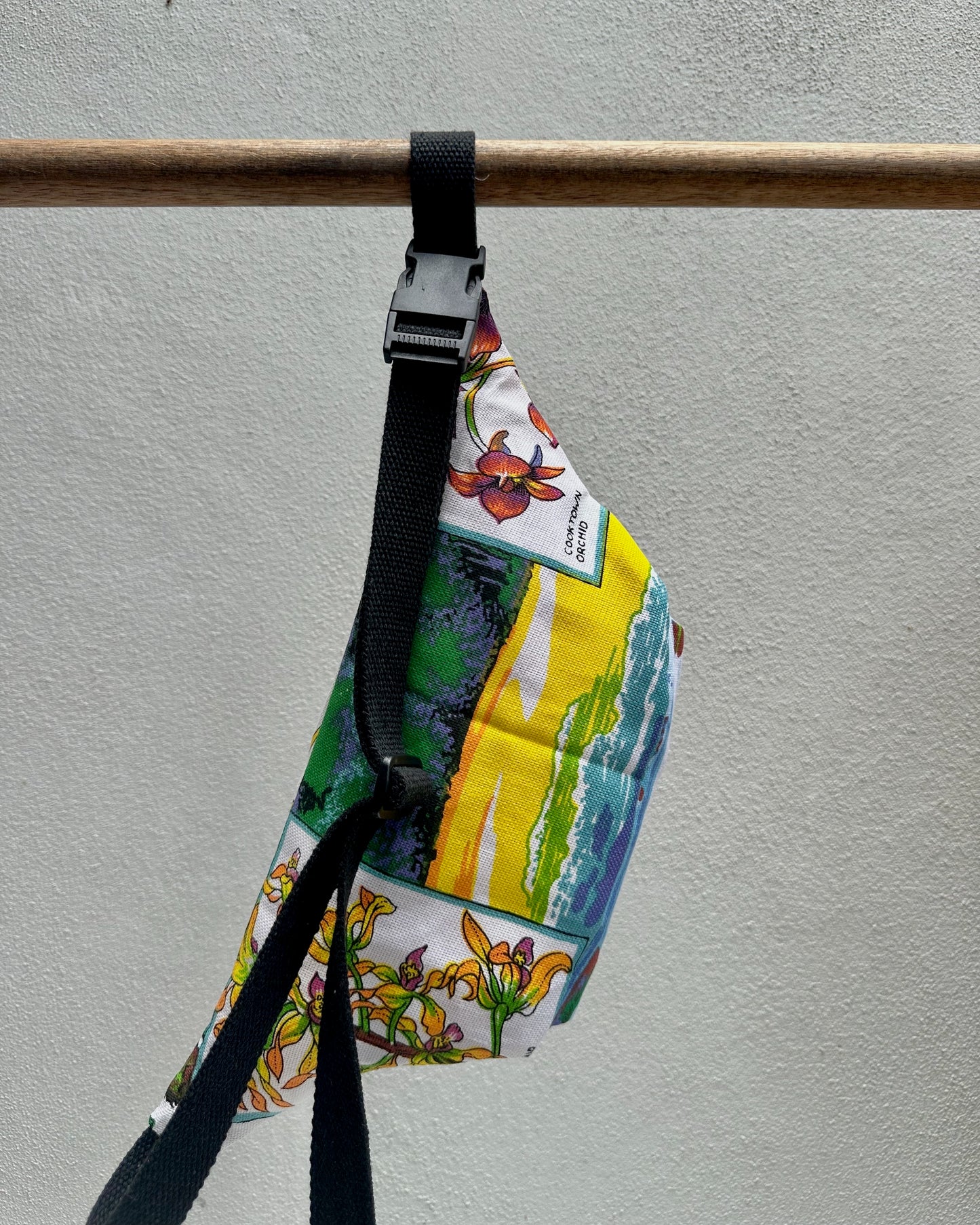 Colorful bag with floral design and black strap hanging on a wooden rod against a gray background