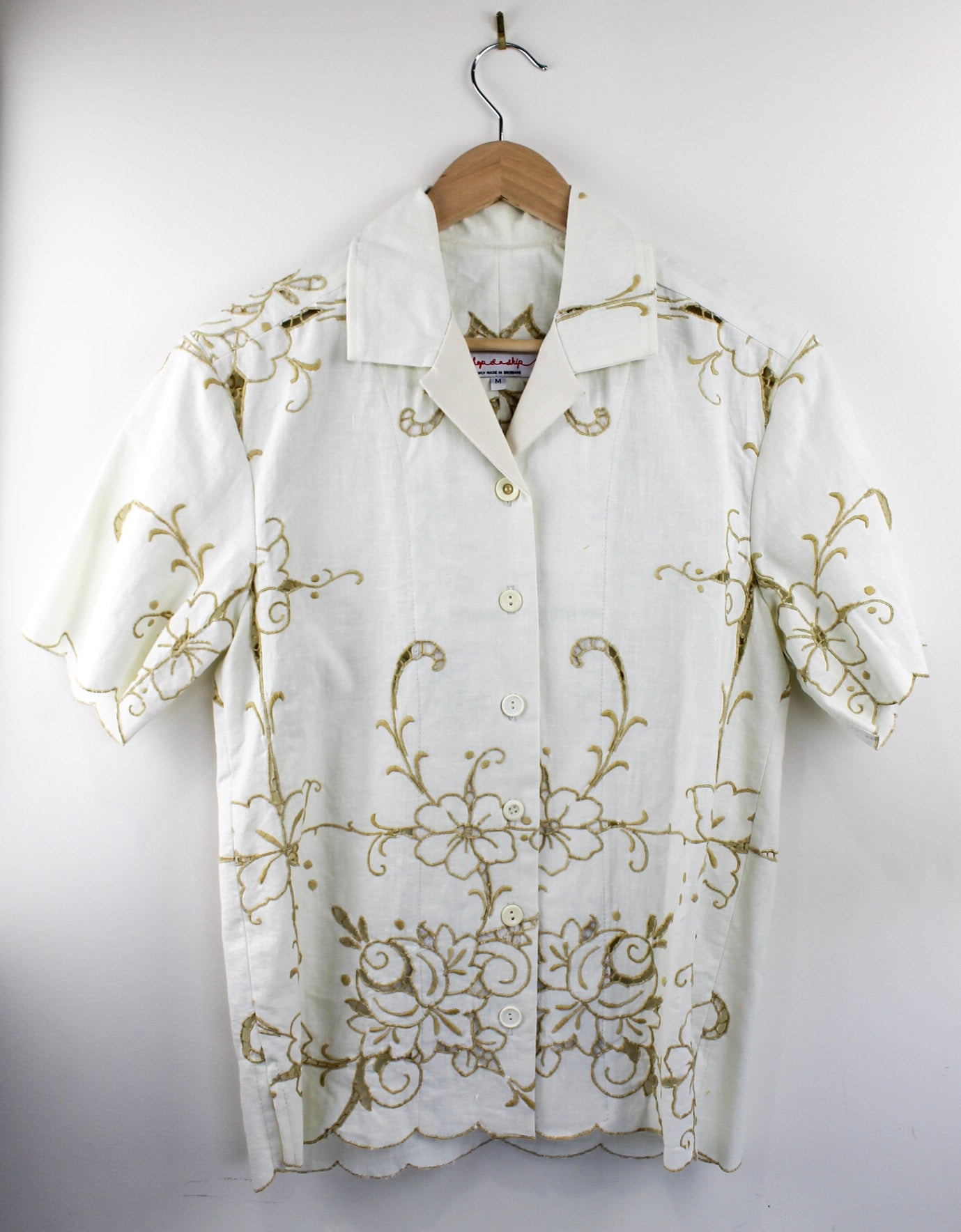 White shirt made from vintage linen with gold embroidery on a wooden hanger against a white background