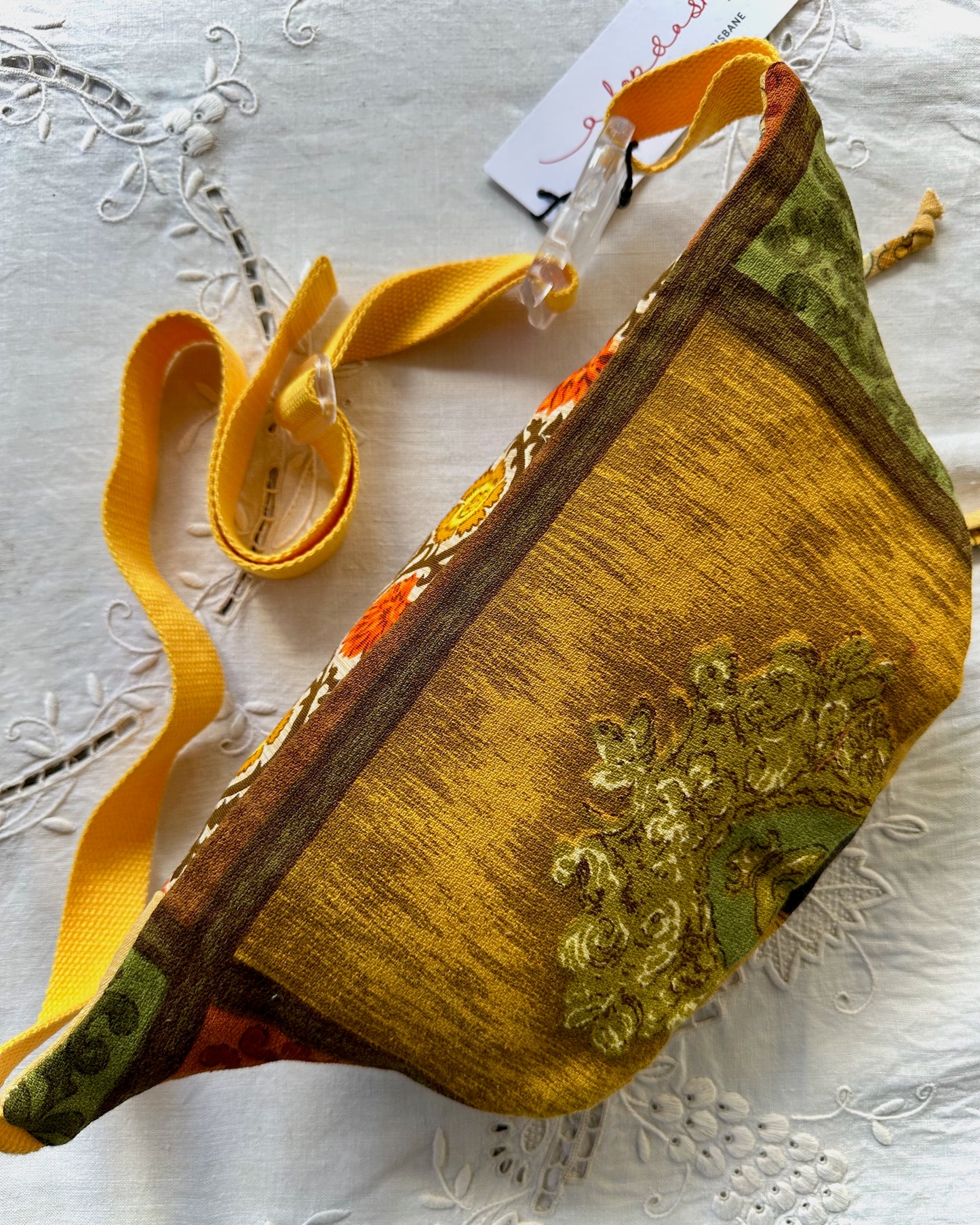 A handmade patchwork fanny pack with a mustard yellow textured body, green patterned fabric, and a yellow webbed strap, laid flat on a white embroidered fabric background.