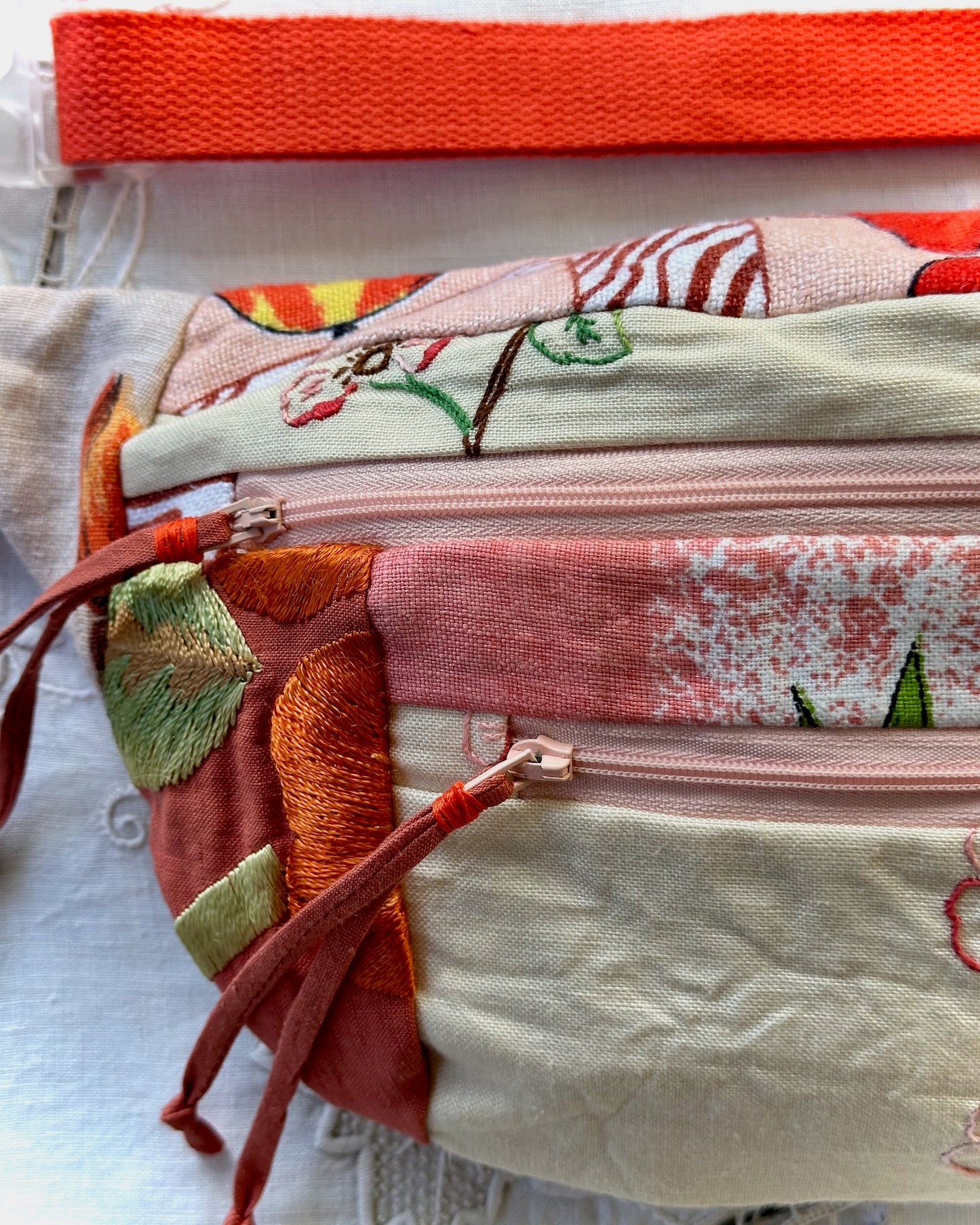 A close-up of a handmade quilted fabric bumbag with a pink zipper, featuring a patchwork design of cream, pink, rust, and green embroidered floral and leaf patterns, with a bright orange-red strap at the top.