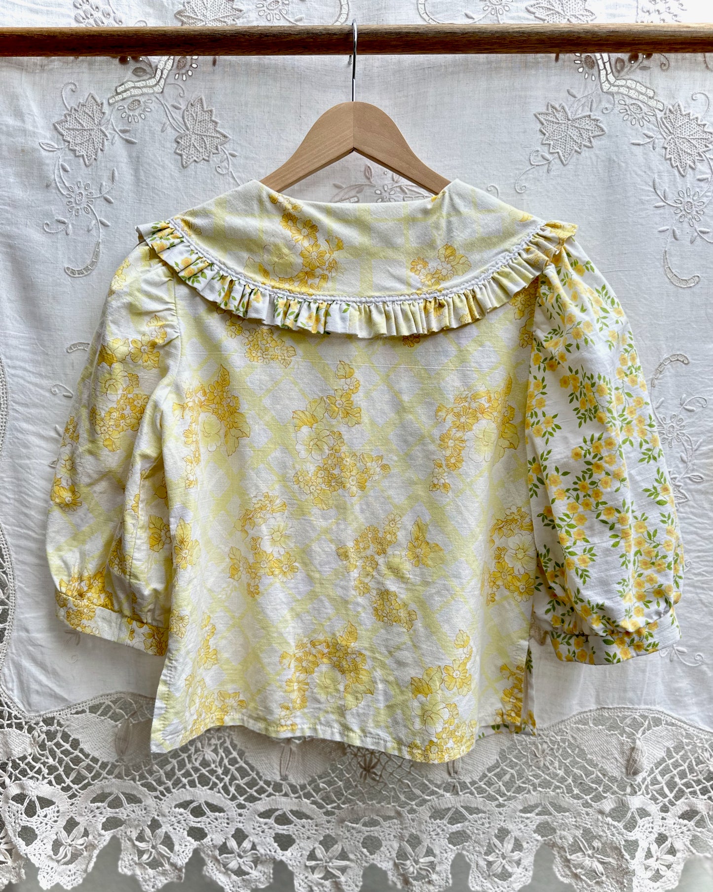 A yellow and white floral print blouse with a ruffled collar and puff sleeves, hanging on a wooden hanger against a white lace curtain backdrop.