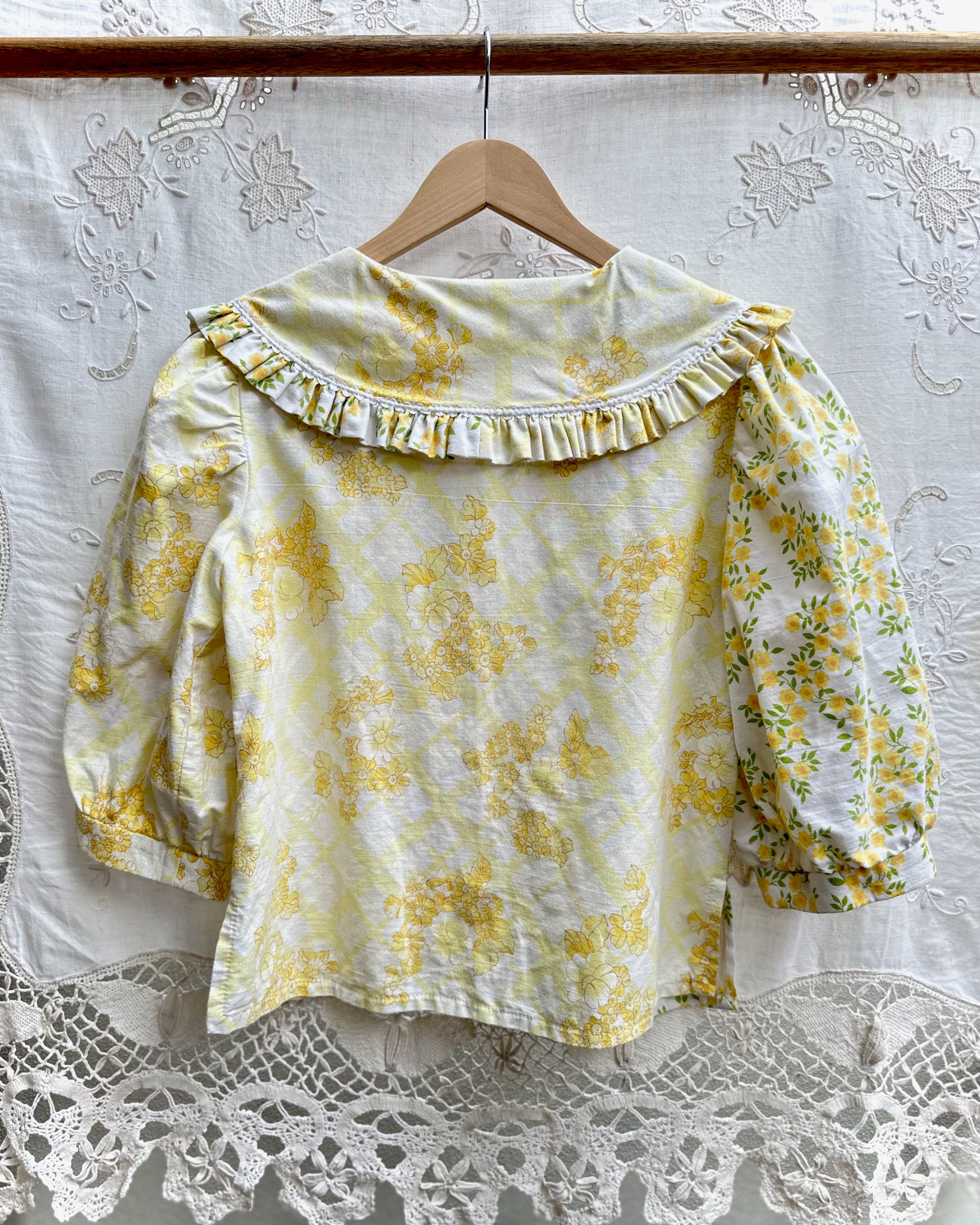 A yellow and white floral print blouse with a ruffled collar and puff sleeves, hanging on a wooden hanger against a white lace curtain backdrop.