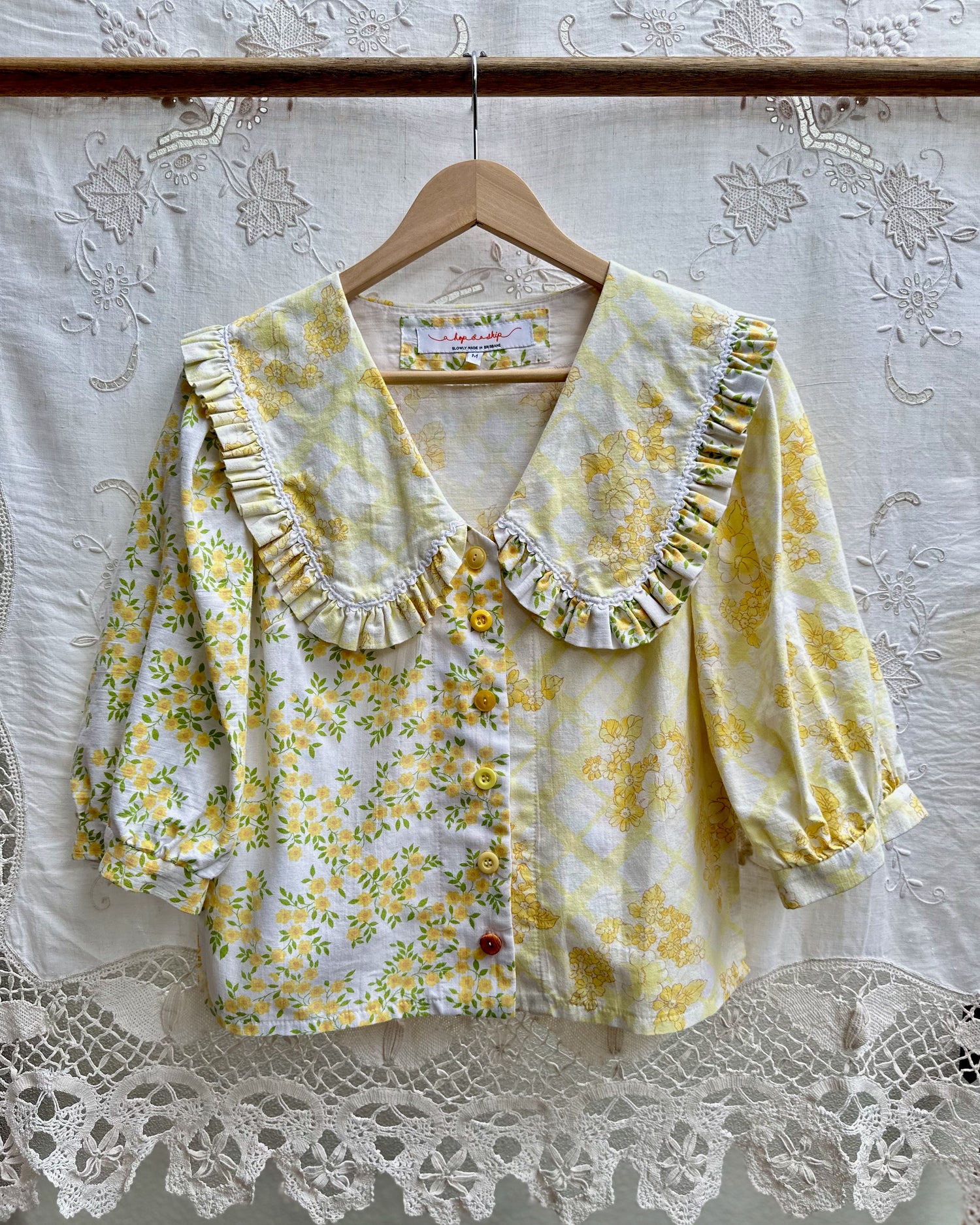 A yellow and white floral print blouse with a large ruffled Peter Pan collar and puff sleeves, hanging on a wooden hanger against a white lace curtain backdrop.
