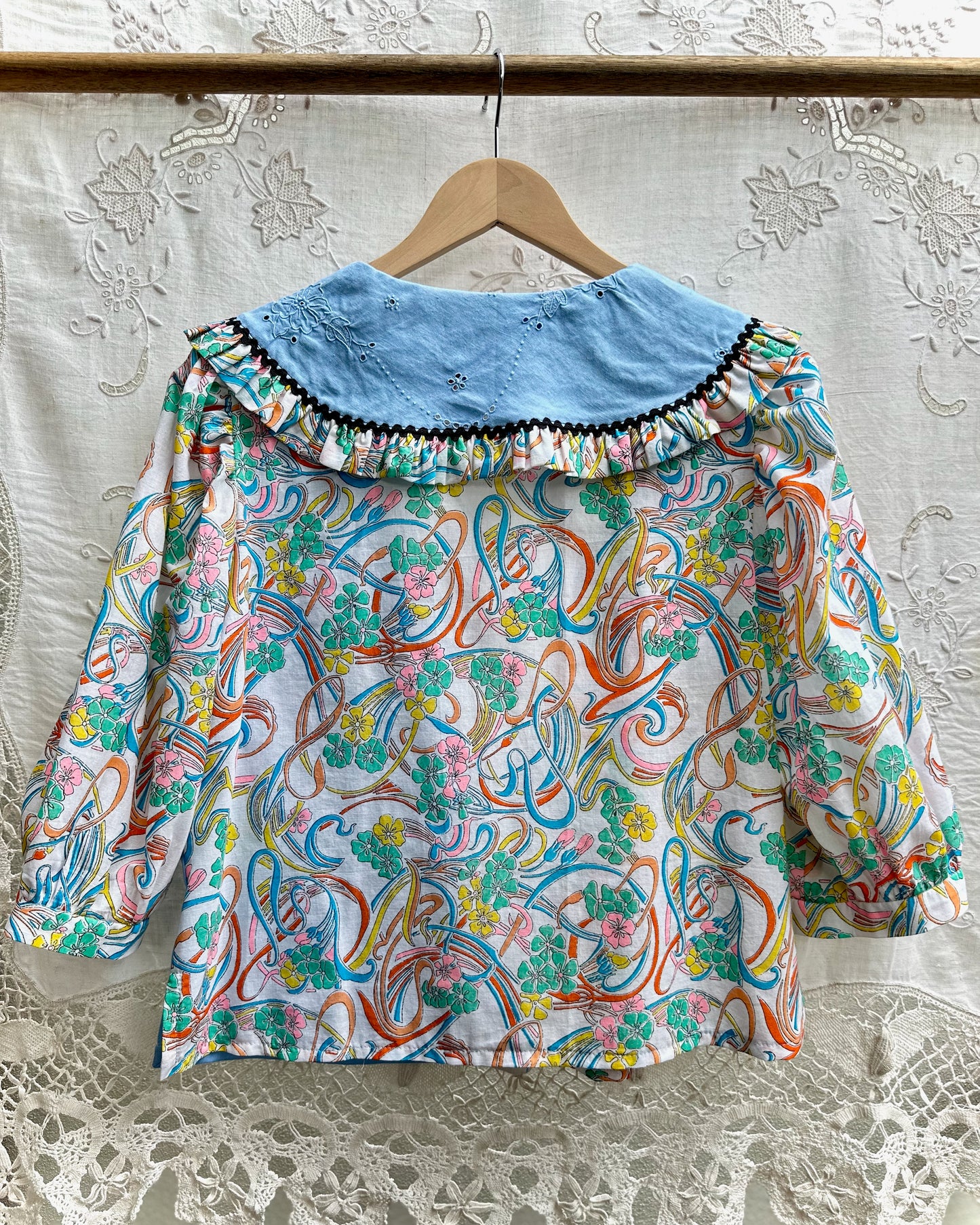 A long-sleeve blouse hanging on a wooden hanger against a white lace curtain backdrop. The blouse features a light blue collar with black trim and a white body covered in a colorful abstract swirl and floral pattern in teal, orange, pink, and yellow.
