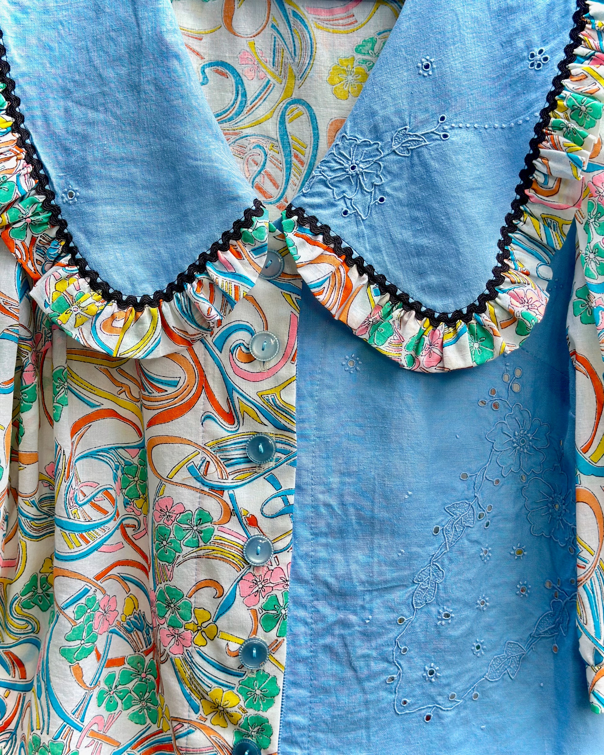 A close-up of a vintage-style blouse featuring a light blue collar with black scalloped trim and embroidered details, layered over a multicolored floral and swirl-patterned blouse.