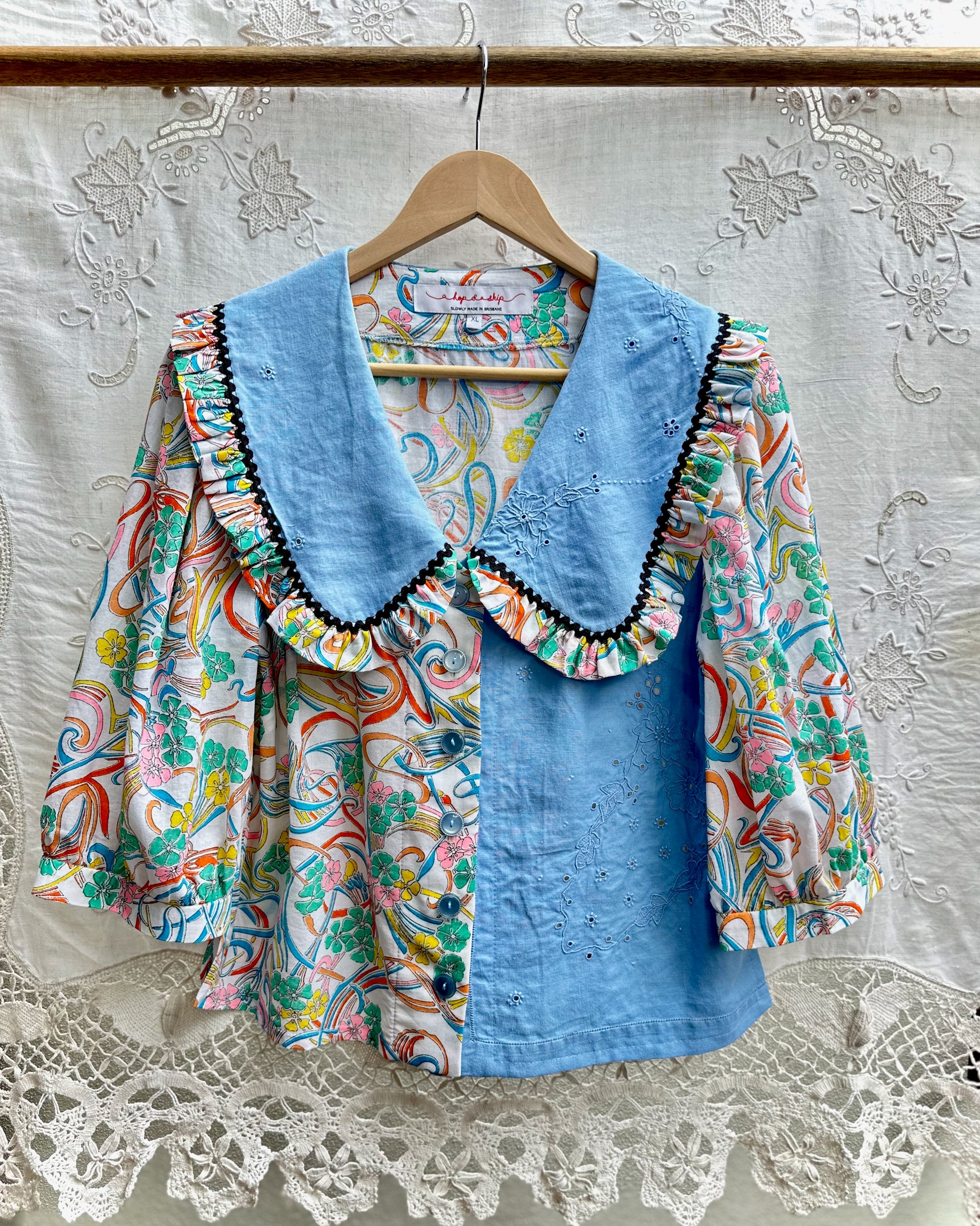 A women's blouse hanging on a wooden hanger against a white embroidered lace curtain. The blouse features a light blue collar and front panel with black trim, paired with a multicolor paisley print on the sleeves and body.