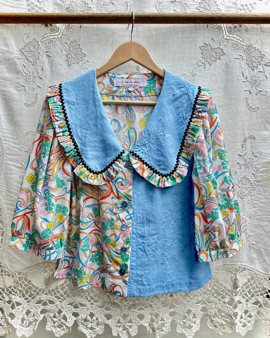 A women's blouse hanging on a wooden hanger against a white embroidered lace curtain. The blouse features a light blue collar and front panel with black trim, paired with a multicolor paisley print on the sleeves and body.
