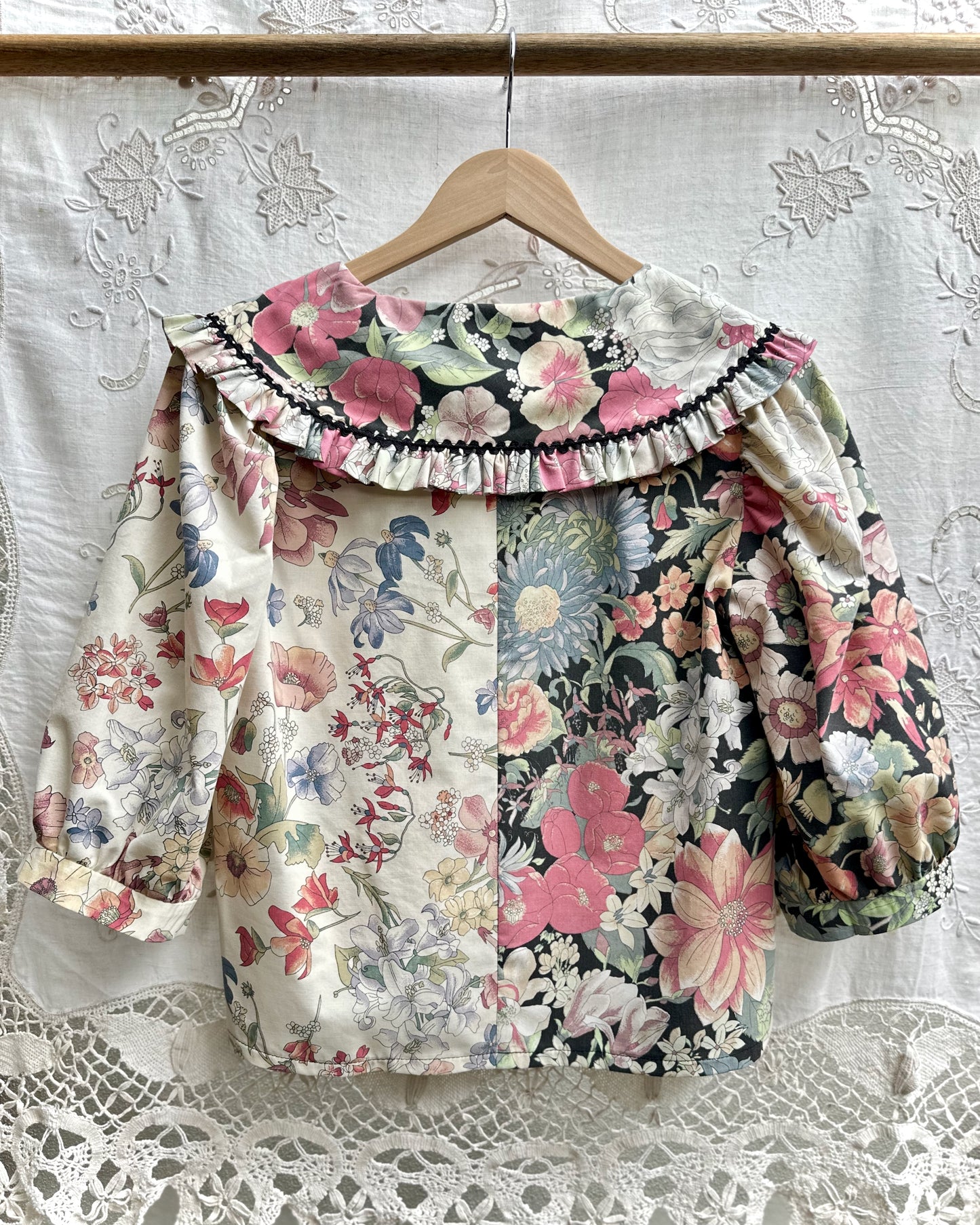 A blouse hanging on a wooden hanger against a white lace curtain. The top has a split floral design: one side is light cream with pink, blue, and green flowers, and the other side is black with pink and blue floral print.