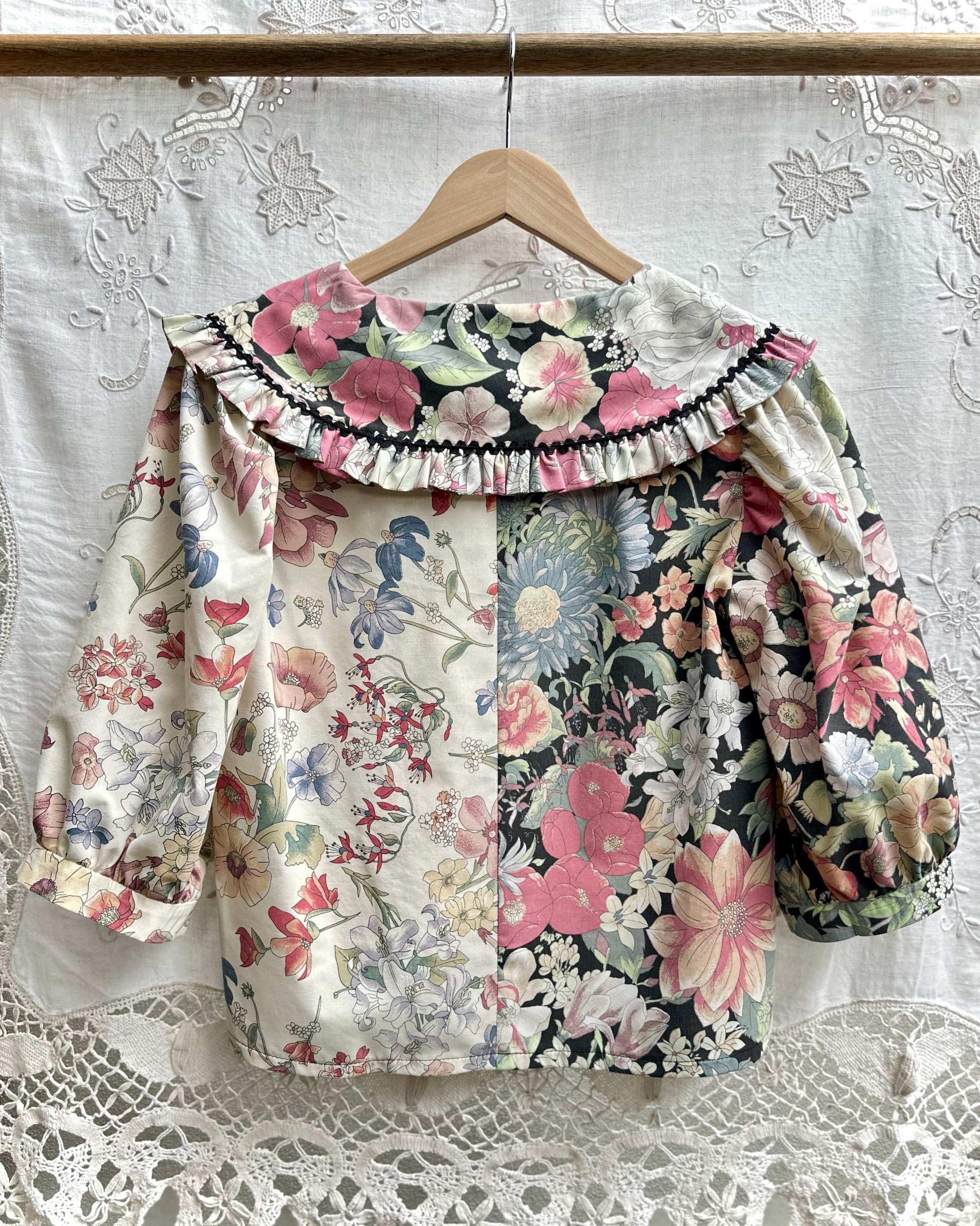 A blouse hanging on a wooden hanger against a white lace curtain. The top has a split floral design: one side is light cream with pink, blue, and green flowers, and the other side is black with pink and blue floral print.
