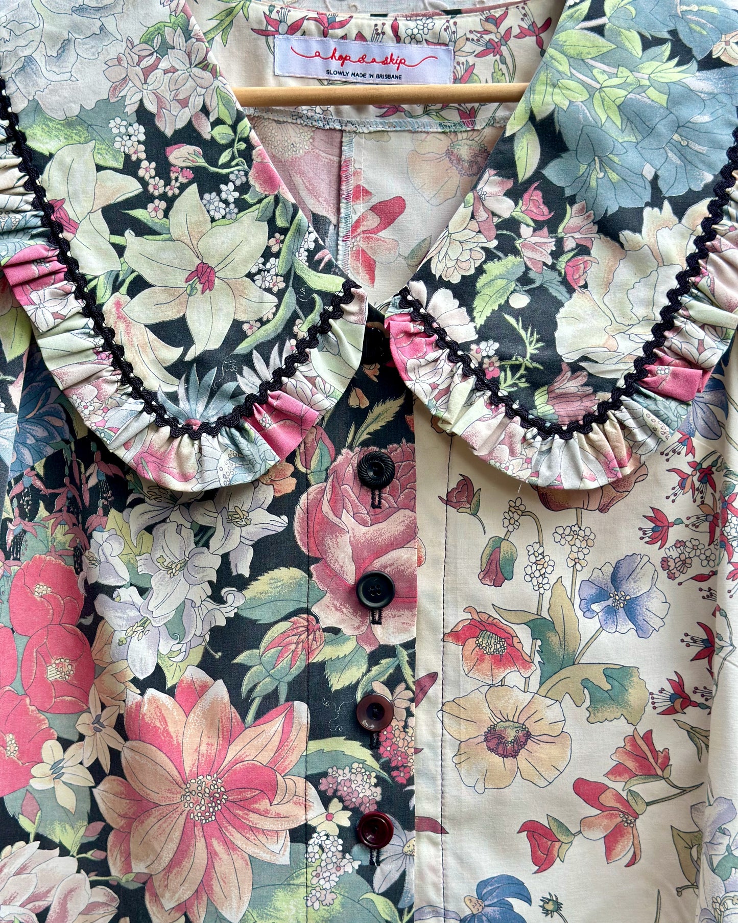 A close-up of a floral button-front blouse with a large ruffled collar. The fabric features a multicolor botanical print with pink, green, blue, and cream tones, accented by black ruffle trim along the collar edges.