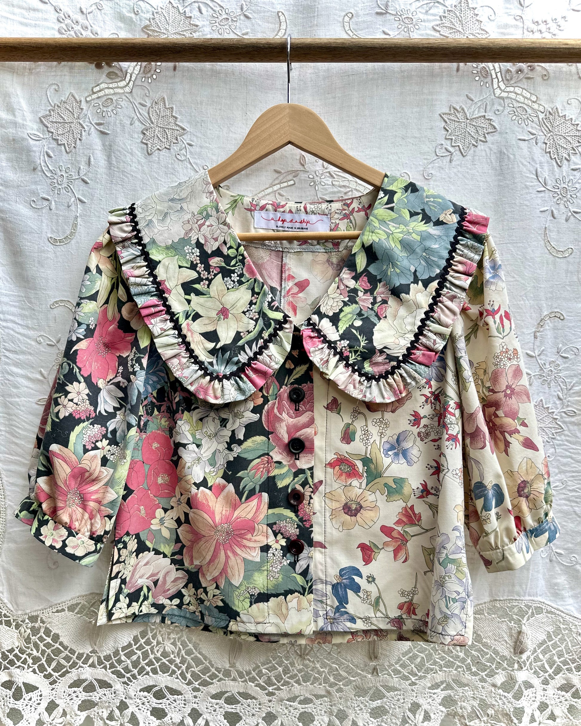 A vintage-style floral blouse with a large ruffled collar, hanging on a wooden hanger against a white lace curtain backdrop.