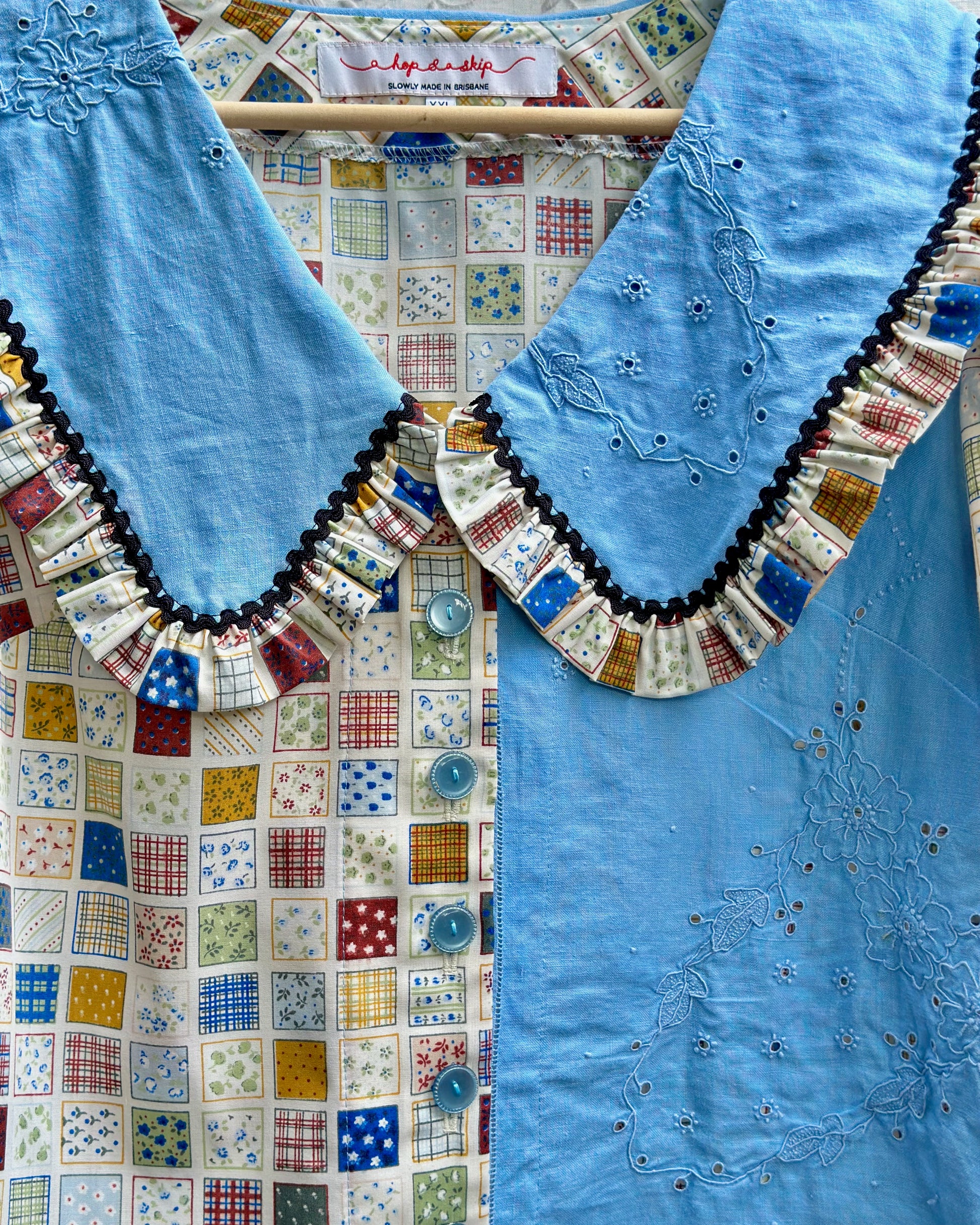 A close-up of a light blue embroidered blouse with a large ruffled collar. The collar is decorated with a patchwork border of small multicolored squares, and the blouse features delicate floral embroidery on the chest.