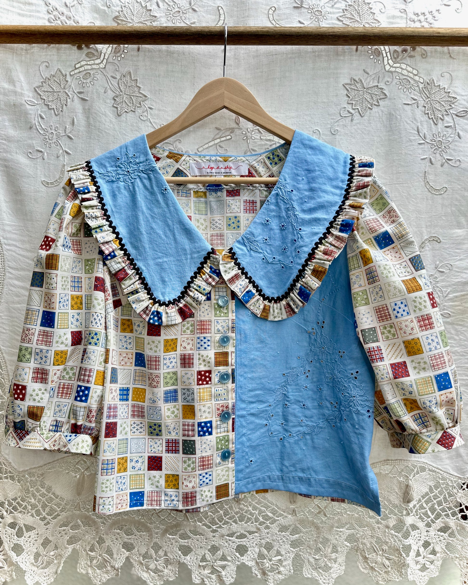 A vintage-style patchwork blouse hanging on a wooden hanger against a white lace curtain backdrop. The blouse features a light blue embroidered bodice and collar, with patchwork-patterned sleeves and front panels in various colorful squares.