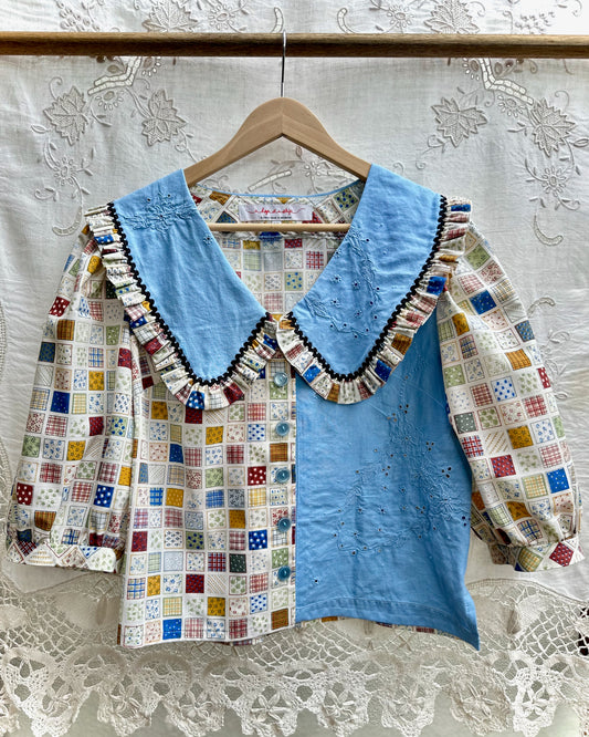 A vintage-style patchwork blouse hanging on a wooden hanger against a white lace curtain backdrop. The blouse features a light blue embroidered bodice and collar, with patchwork-patterned sleeves and front panels in various colorful squares.