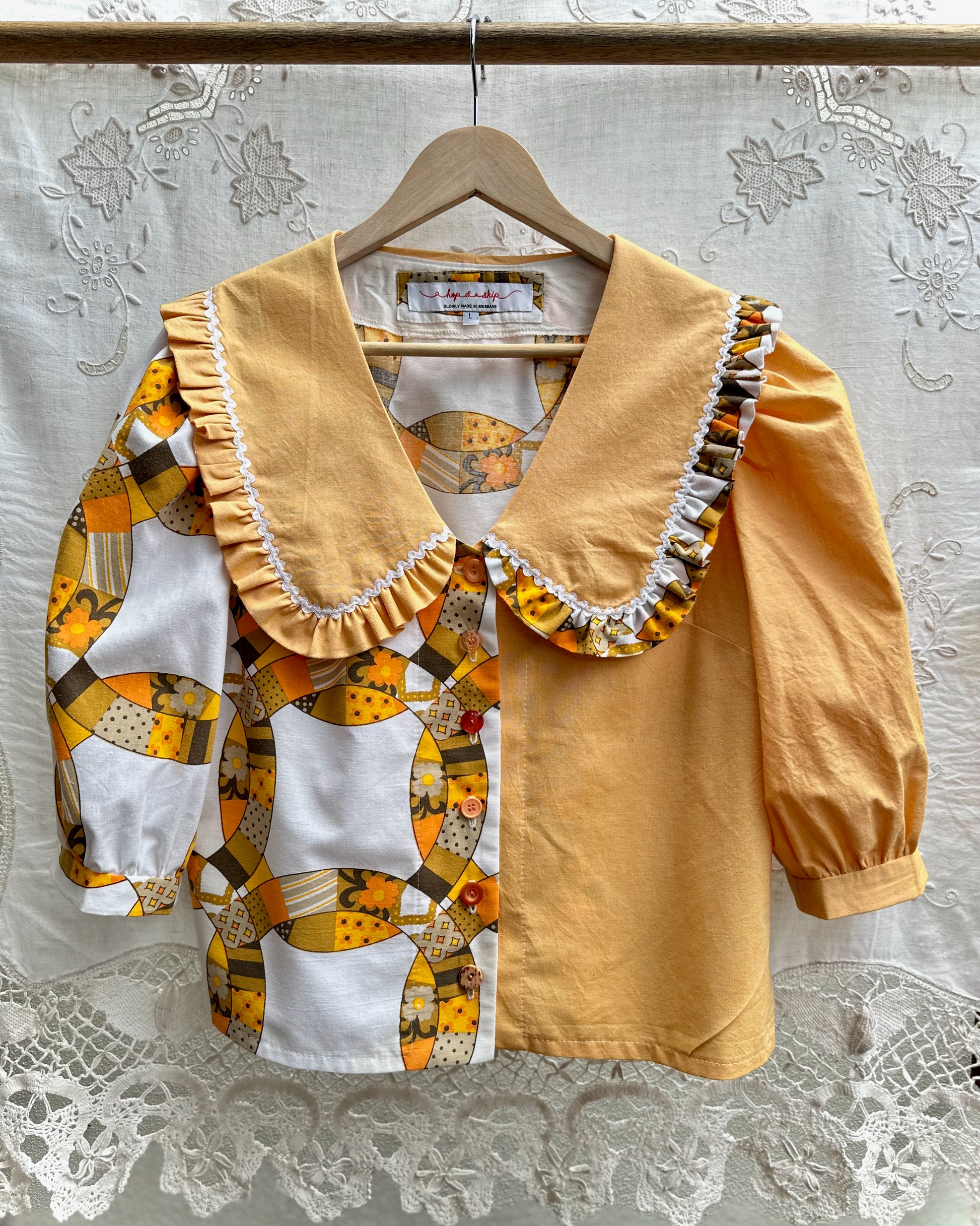 A children's blouse hanging on a wooden hanger against a white lace curtain backdrop. The top has a solid mustard yellow right side with a large ruffled collar, and a left side with a patchwork print of yellow, white, and brown geometric shapes.