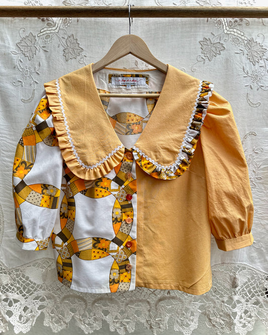A children's blouse hanging on a wooden hanger against a white lace curtain backdrop. The top has a solid mustard yellow right side with a large ruffled collar, and a left side with a patchwork print of yellow, white, and brown geometric shapes.