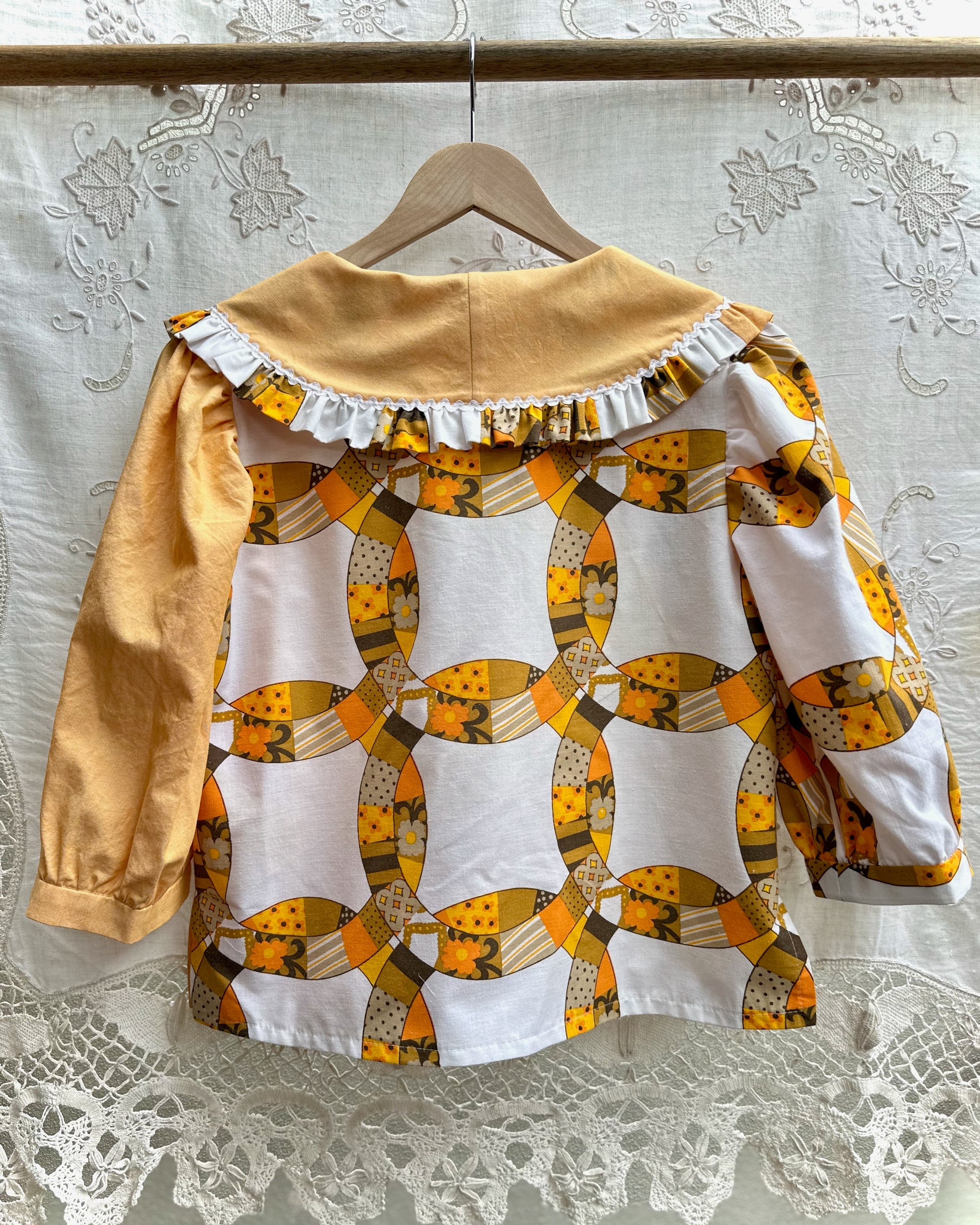 A long-sleeve blouse hanging on a wooden hanger against a white lace curtain backdrop. The back view shows a large-scale geometric patchwork pattern in yellow, orange, white, and brown, with a solid mustard yellow ruffled collar and one solid mustard yellow sleeve.