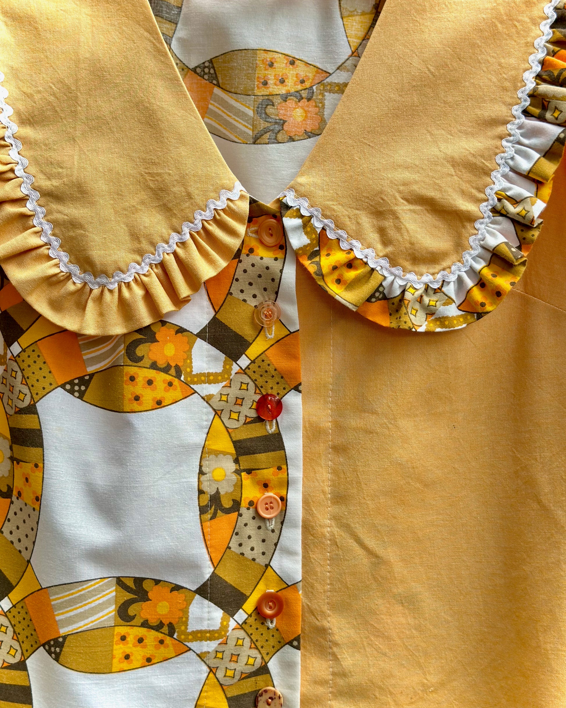 Close-up of a garment featuring a solid mustard yellow collar with white ruffle trim, paired with a patchwork-style patterned section in yellow, white, and brown.