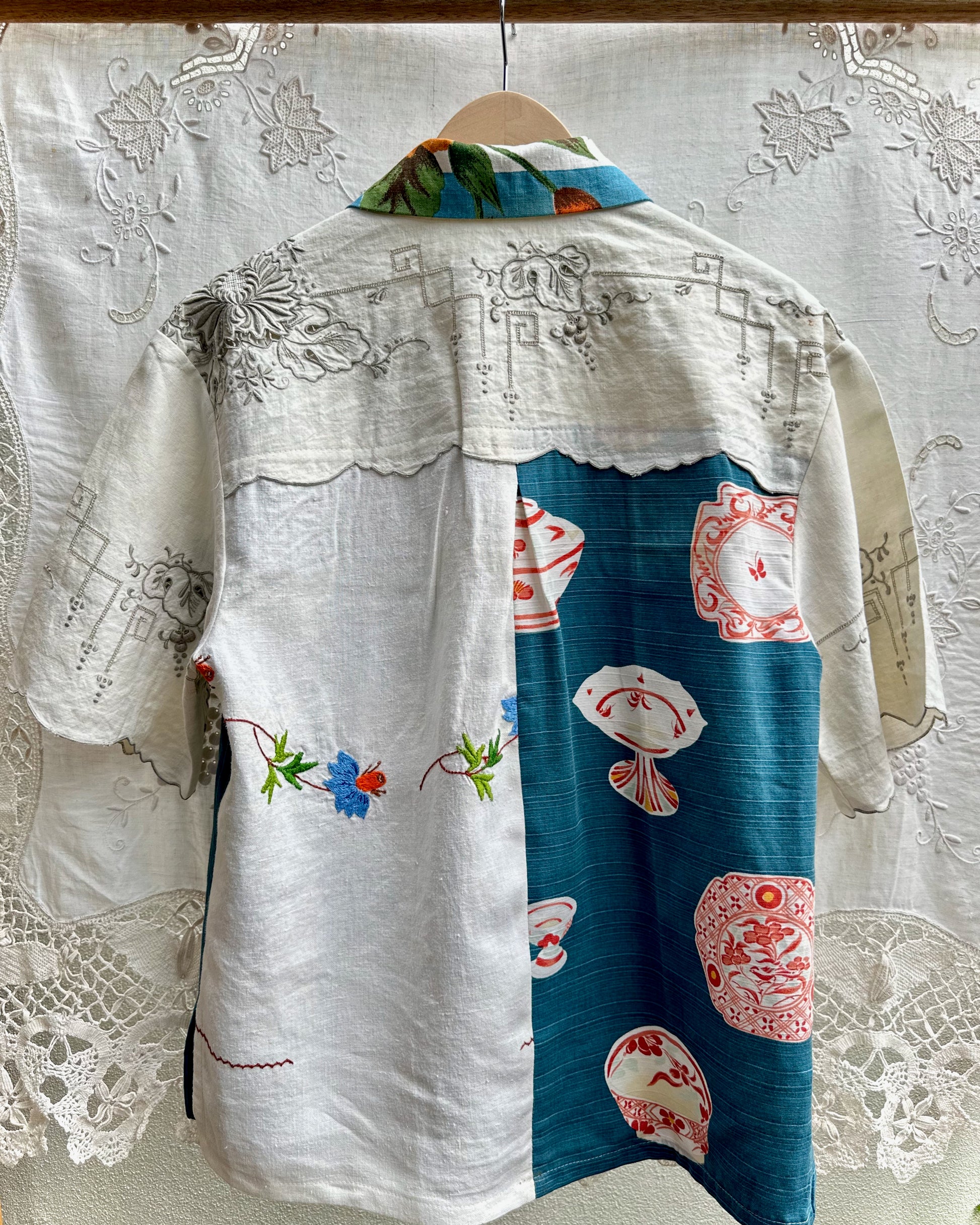 A short-sleeve button-up shirt hanging on a wooden hanger against a white lace curtain backdrop. The shirt features a split design: one side is white with black floral embroidery and small blue, red, and green floral accents, while the other side is teal blue with pink and white teacup patterns.