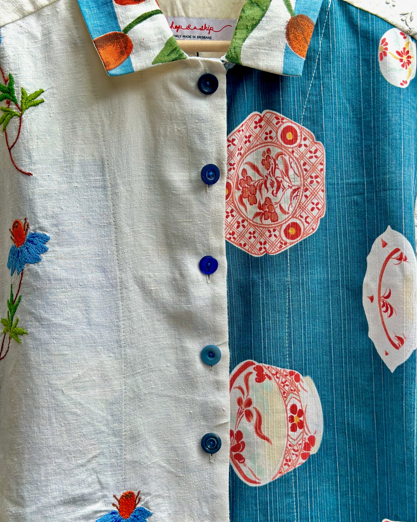 A close-up of a button-front shirt with a split design: one side is off-white with blue floral embroidery, and the other side is teal-blue with red and white decorative motifs. The collar also shows a multicolor print with orange and green details.