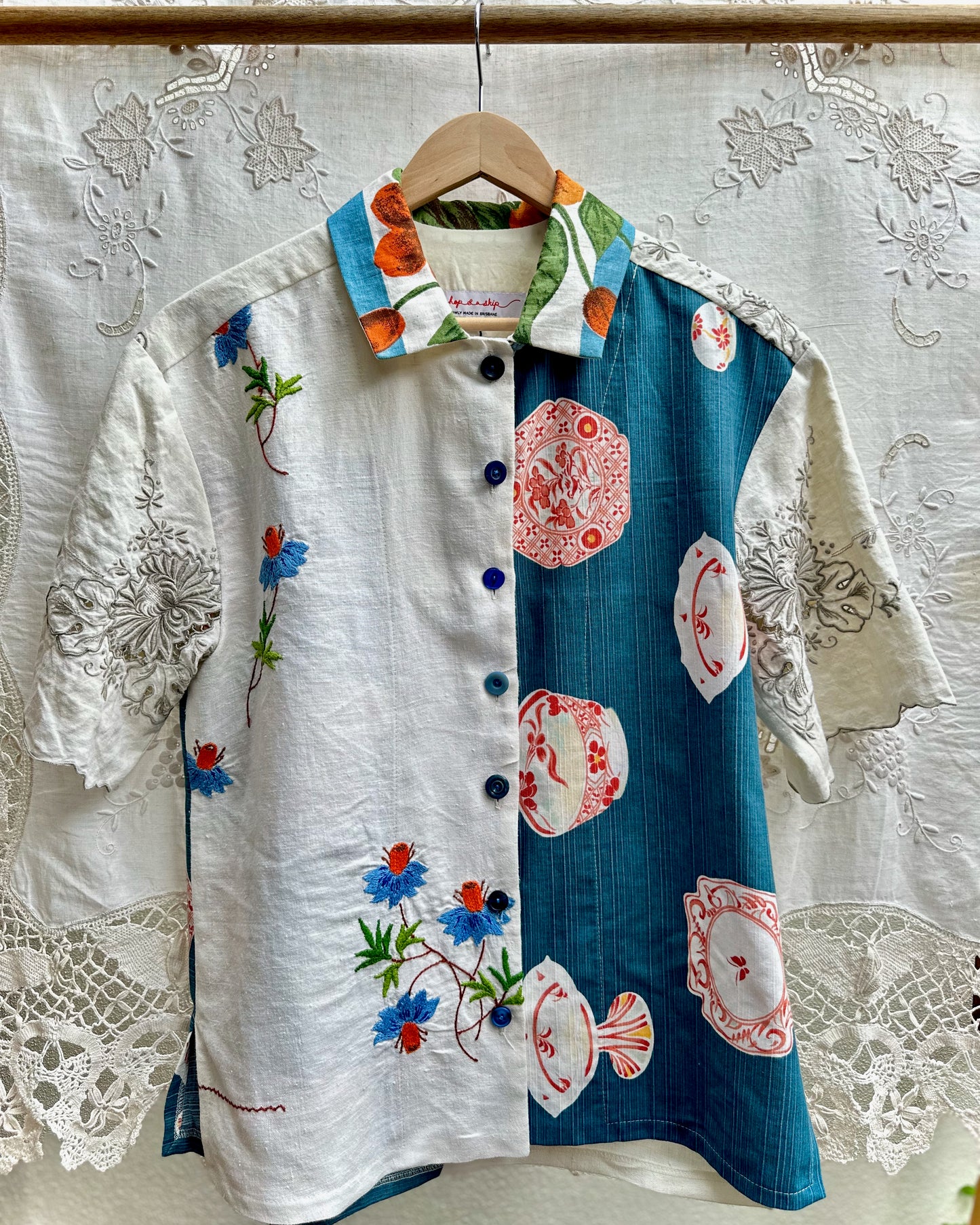A short-sleeve button-up shirt hanging on a wooden hanger against a white lace curtain backdrop. The shirt has a split design: one half is white with blue and red floral embroidery, and the other half is teal blue with pink and white teacup patterns.