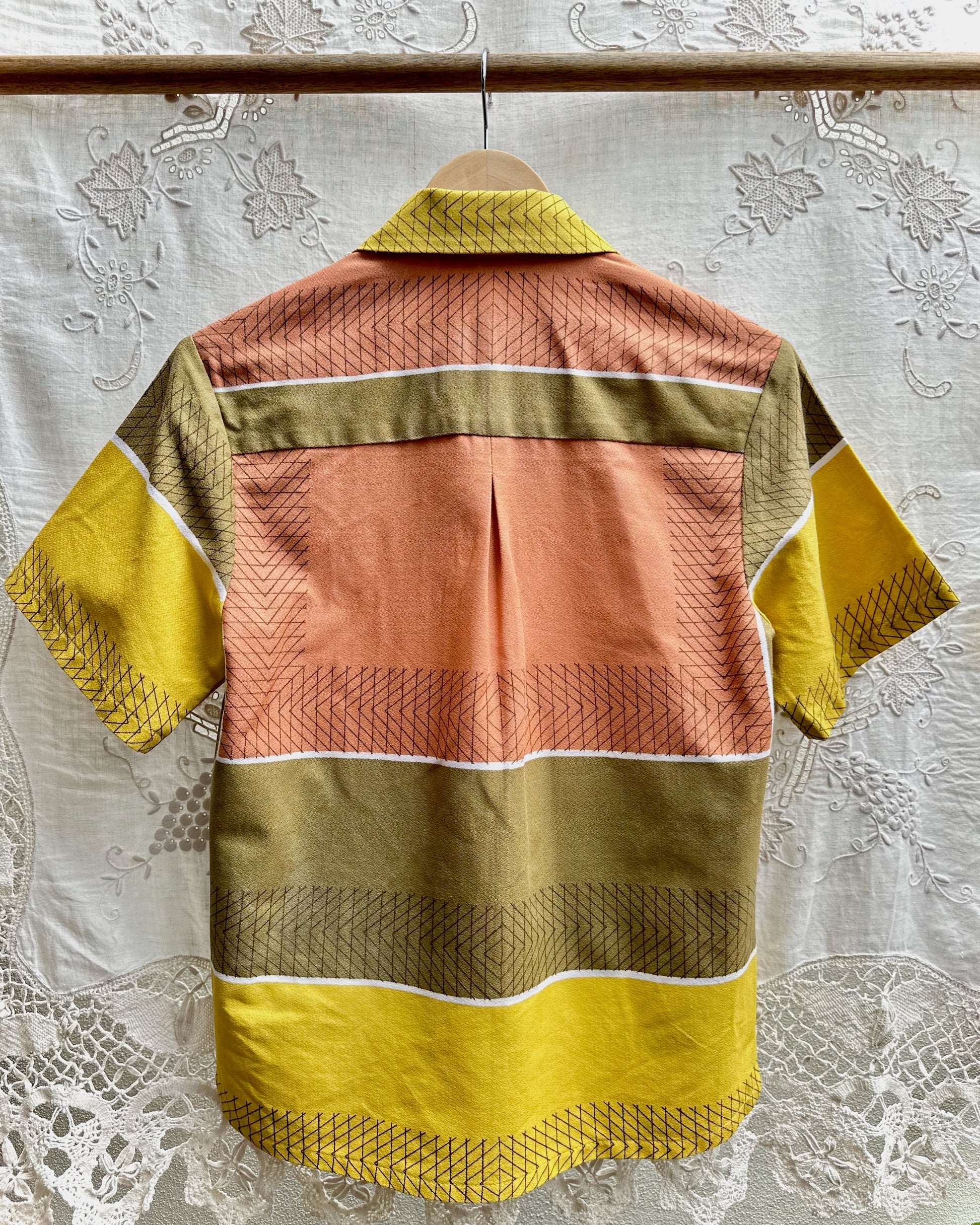 A short-sleeve button-up shirt with a retro striped pattern in yellow, peach, olive green, and brown, hanging on a wooden hanger against a white lace curtain background.