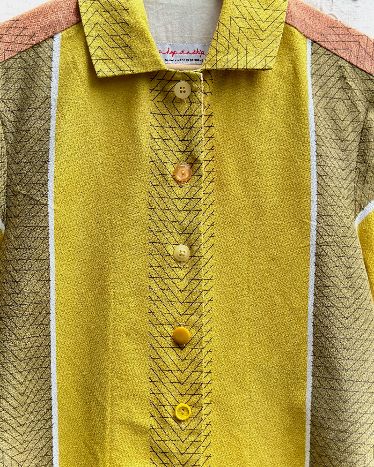 A close-up of a vintage yellow short-sleeve button-up shirt with a geometric patterned collar and cuffs, featuring a central vertical stripe with yellow buttons and peach-colored shoulder panels.