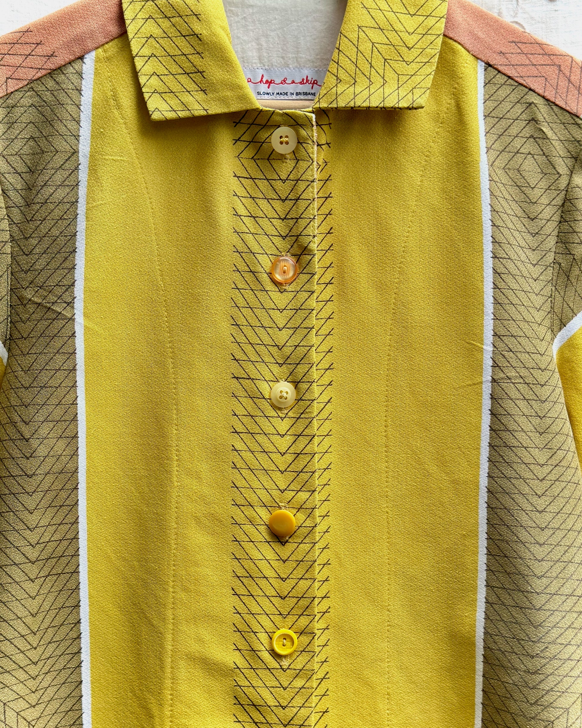 A close-up of a vintage yellow short-sleeve button-up shirt with a geometric patterned collar and cuffs, featuring a central vertical stripe with yellow buttons and peach-colored shoulder panels.