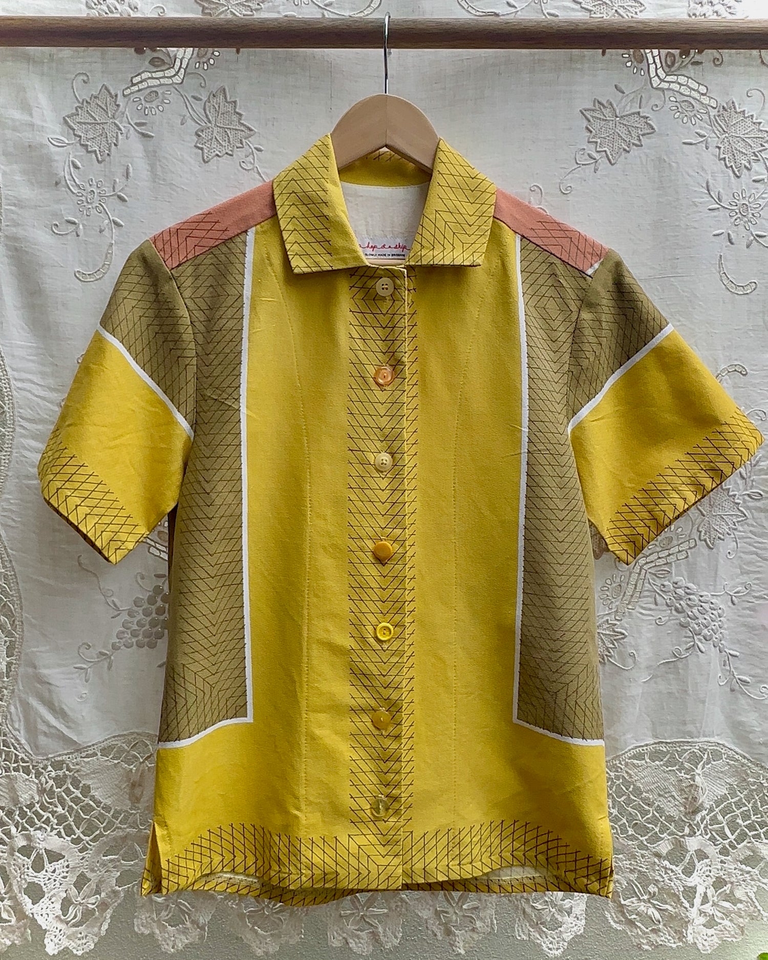 A short-sleeve button-up shirt hanging on a wooden hanger against a white lace curtain backdrop. The shirt features a bright yellow center panel with a button front, contrasted by textured tan side panels and peach-pink shoulder yokes with white piping details.
