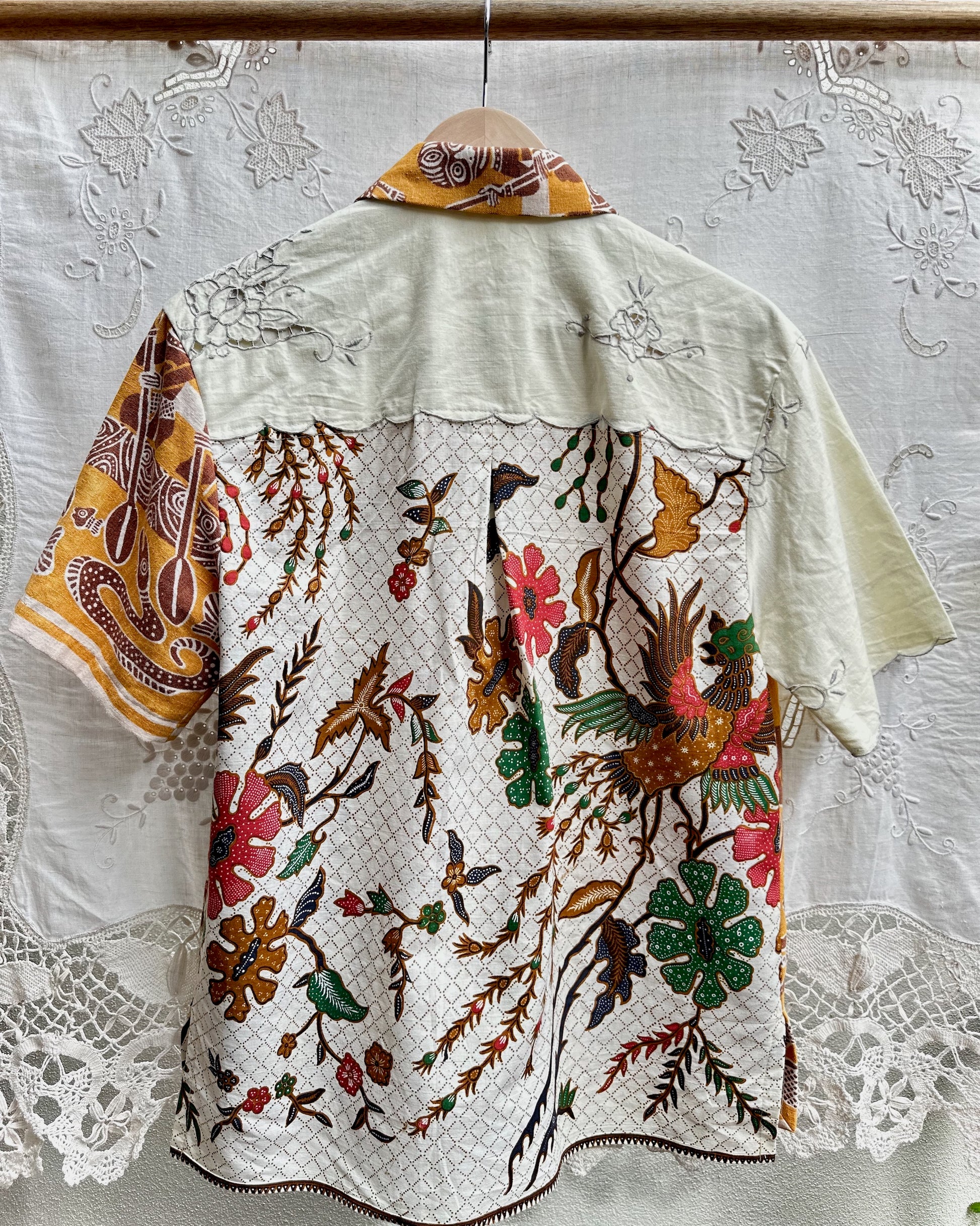 A short-sleeve button-up shirt with a cream-colored base and an all-over batik-style print of birds, flowers, and foliage. The shirt is shown hanging on a wooden hanger against a white lace curtain backdrop.
