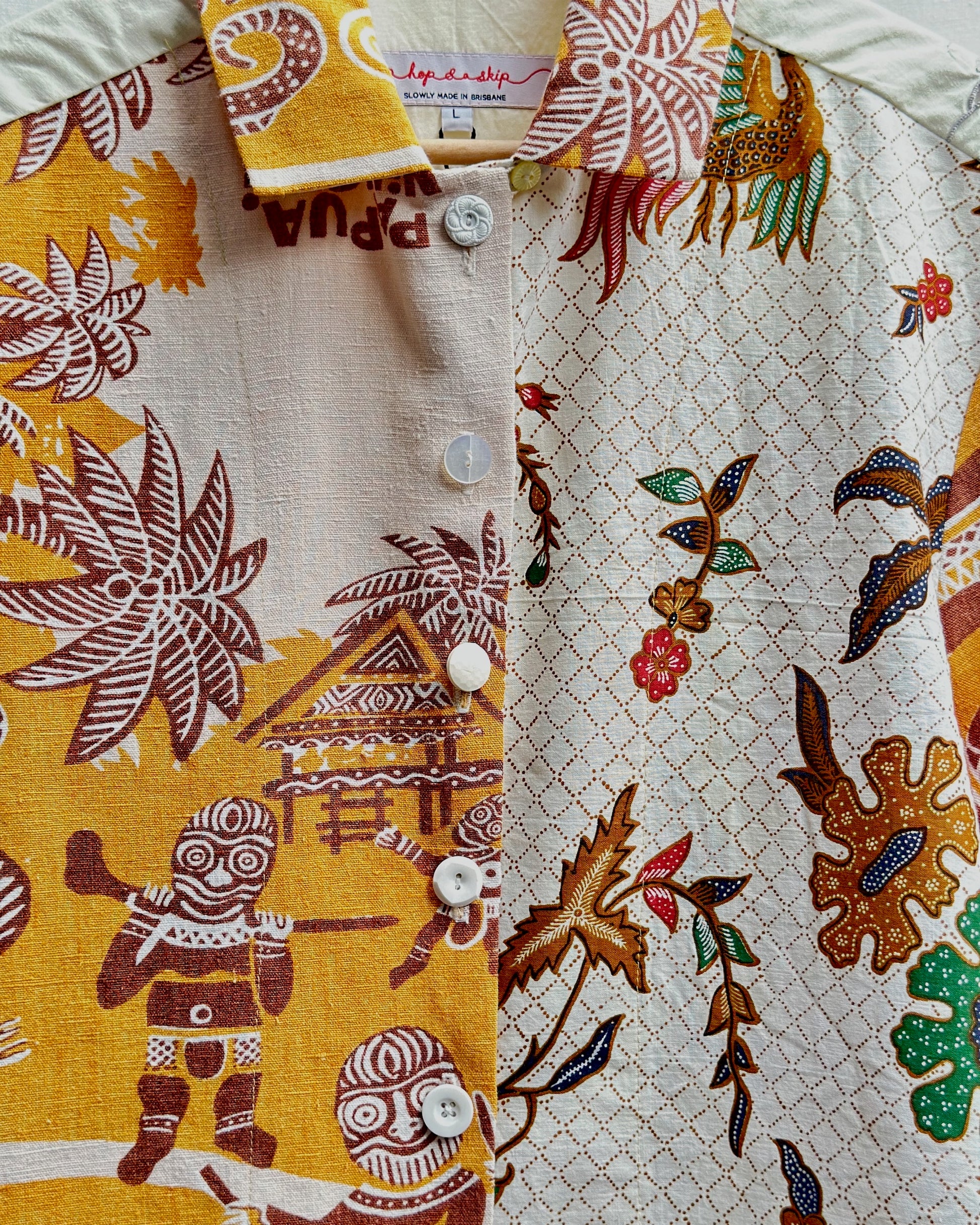 A close-up of a short-sleeve button-up shirt featuring a split design: one side has a mustard yellow background with brown tribal figures and motifs, and the other side has a cream background with a brown diamond lattice pattern and multicolored floral details.