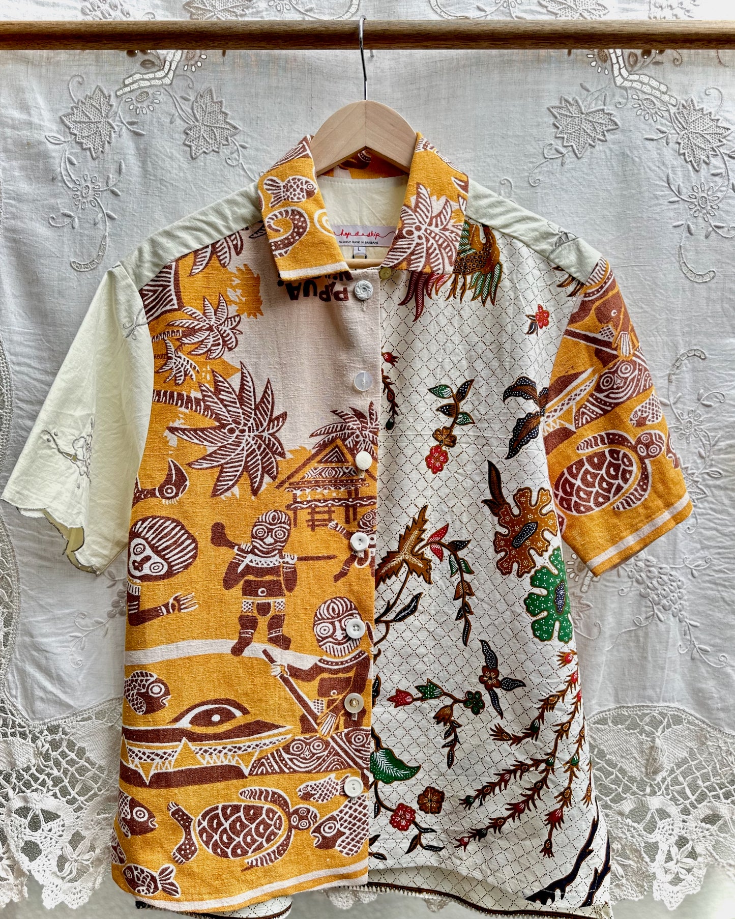 A short-sleeve button-up shirt hanging on a wooden hanger against a white lace curtain backdrop. The shirt features a split design: one side is mustard yellow with brown tribal-style illustrations, and the other side is off-white with a geometric pattern and botanical motifs in brown, green, and red.