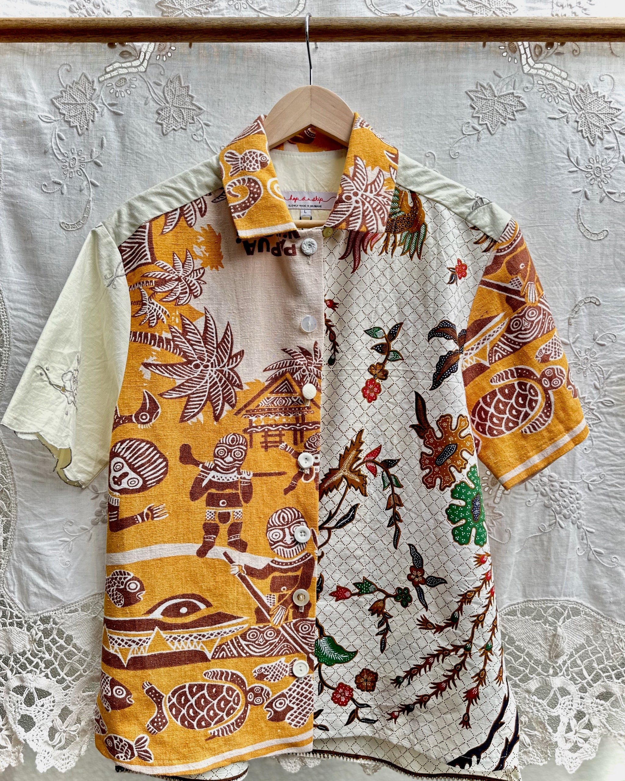 A short-sleeve button-up shirt hanging on a wooden hanger against a white lace curtain backdrop. The shirt features a split design: one side is mustard yellow with brown tribal-style illustrations, and the other side is off-white with a geometric pattern and botanical motifs in brown, green, and red.