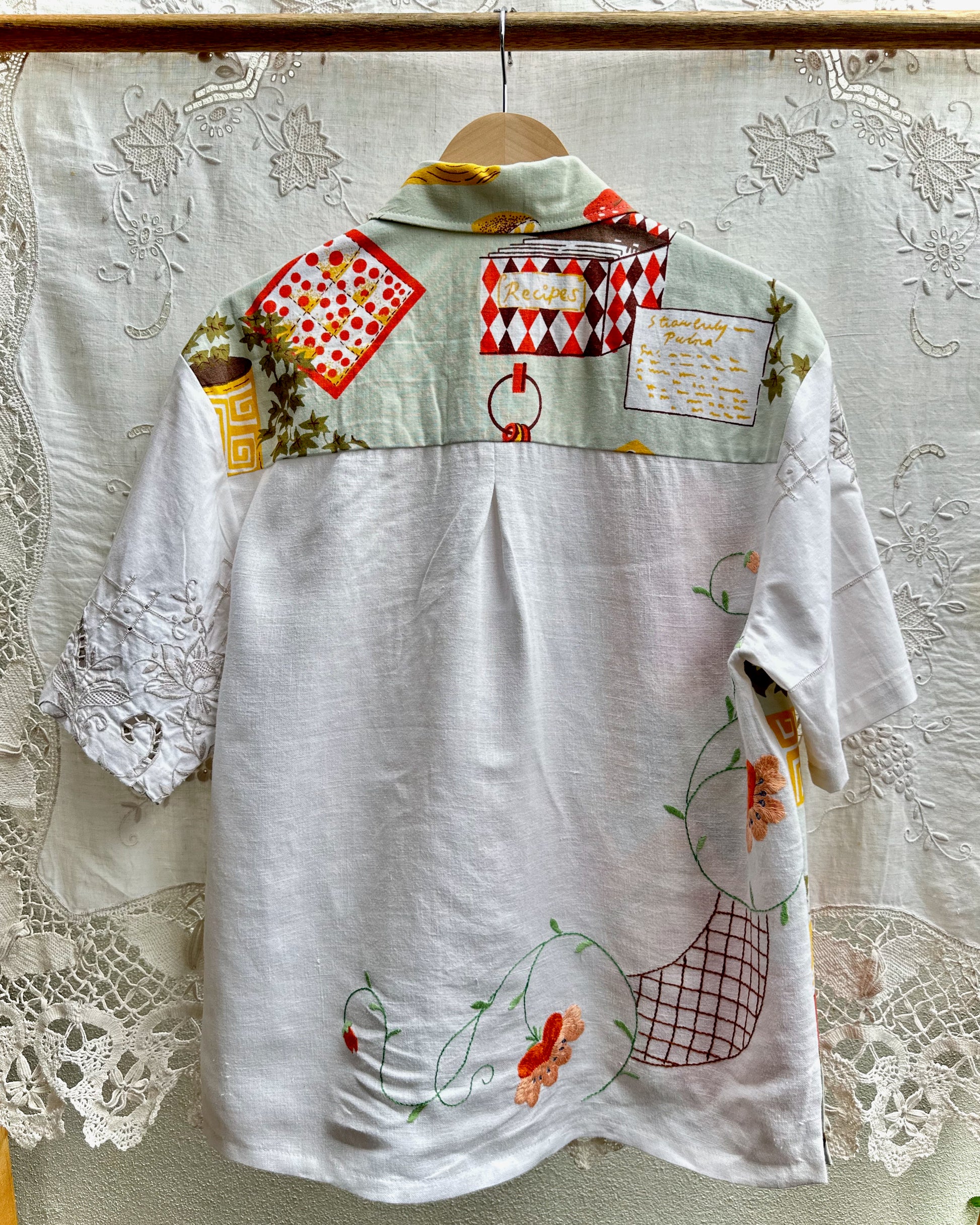 A white short-sleeve button-up shirt with a highly decorative, multicolored patchwork-style print on the upper back and shoulders, hanging on a wooden hanger against a white lace curtain backdrop.