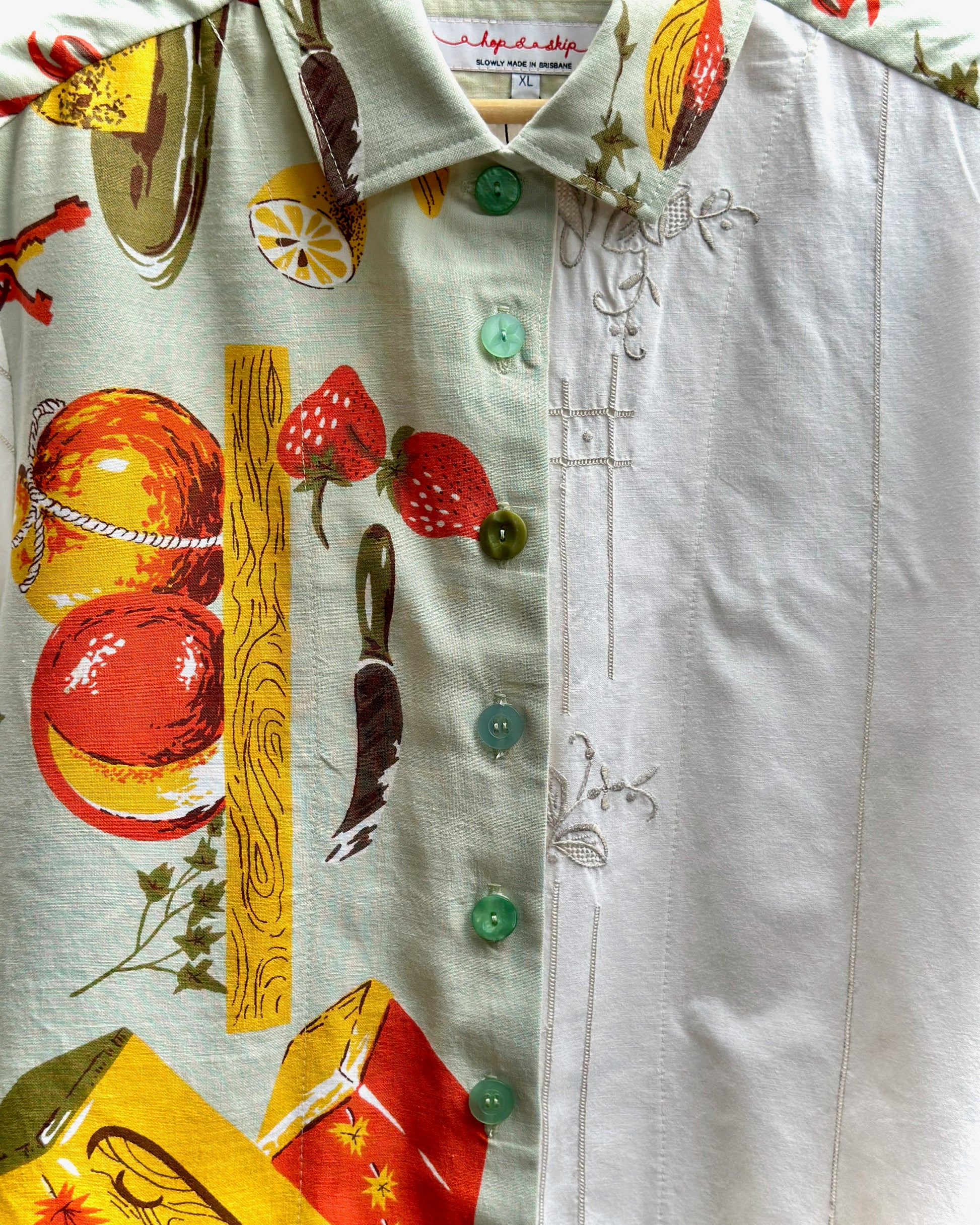 A close-up of a white button-up shirt featuring a split design: one side has a colorful fruit and citrus print with green buttons, and the other side has delicate gray floral embroidery.