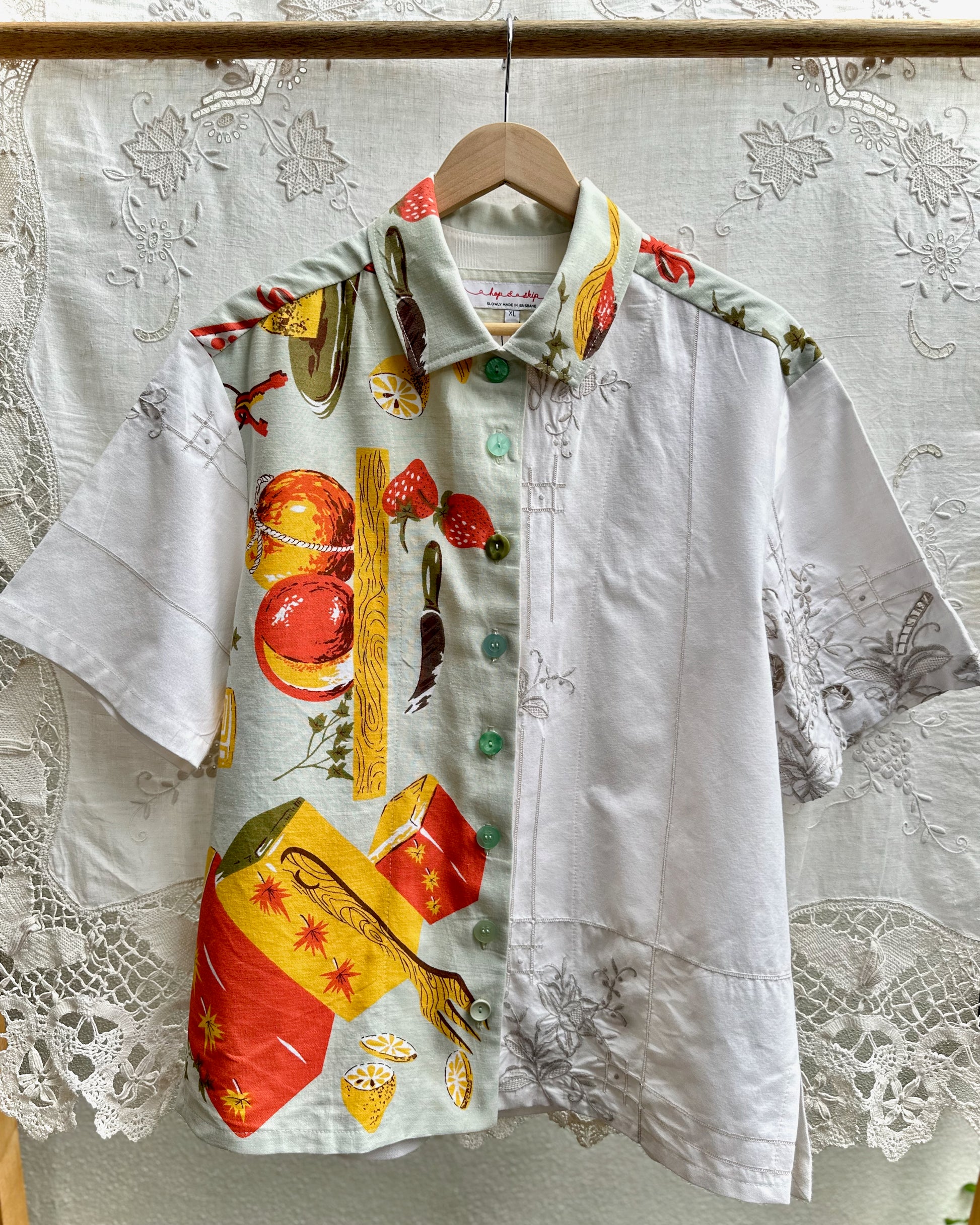 A short-sleeve button-up shirt hanging on a wooden hanger against a white lace curtain backdrop. The shirt has a split design: one side is solid white with subtle floral embroidery, while the other side features a colorful print of illustrated fruit, cigars, and cigars with red, yellow, green, and brown tones.