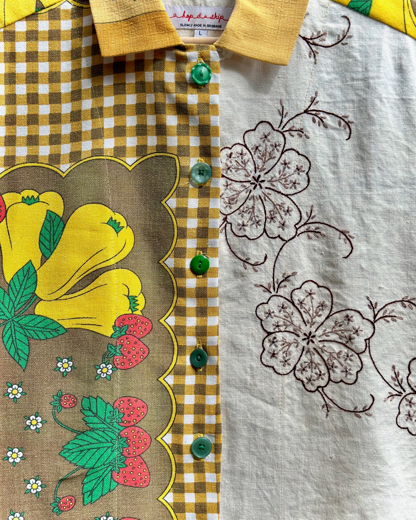 A close-up of a collared shirt featuring a split design: one side has a brown and yellow gingham pattern with a scalloped border and colorful yellow, green, and red floral print, while the other side has a white linen-like fabric with brown floral embroidery. Green buttons are visible down the center.