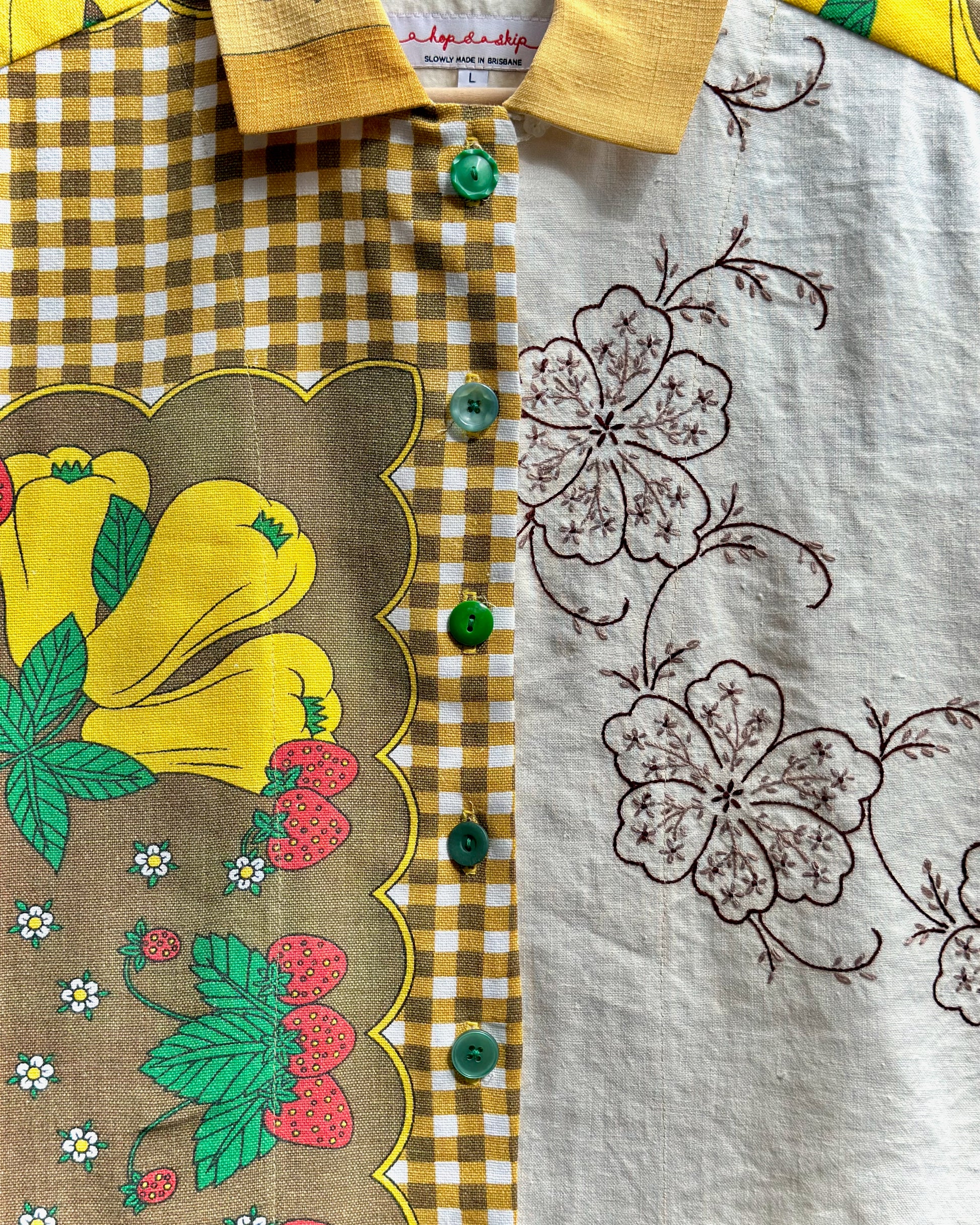 A close-up of a collared shirt featuring a split design: one side has a brown and yellow gingham pattern with a scalloped border and colorful yellow, green, and red floral print, while the other side has a white linen-like fabric with brown floral embroidery. Green buttons are visible down the center.