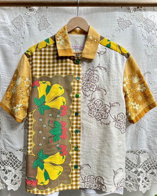 A short-sleeve button-up shirt hanging on a wooden hanger against a white lace curtain backdrop. The shirt features a patchwork design with a yellow gingham panel, a brown panel with large yellow hibiscus flowers and green leaves, and a white panel with black floral line drawings.