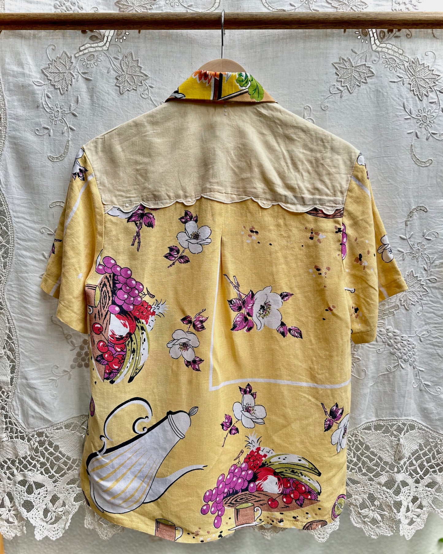 A yellow short-sleeve button-up shirt with a colorful floral and fruit print, hanging on a wooden hanger against a white lace curtain background.