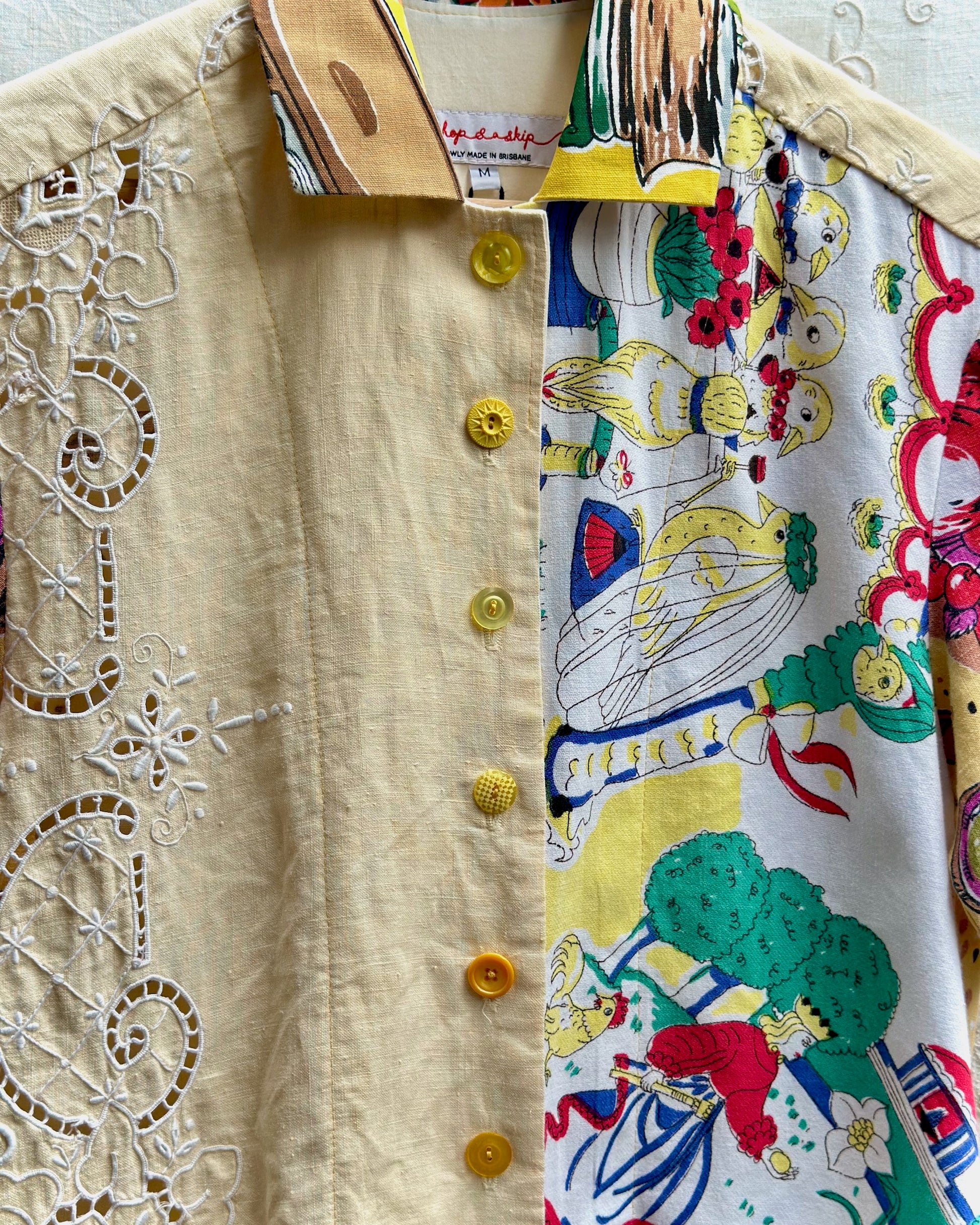 A close-up of a shirt with a split design: one half is light beige with white floral embroidery and yellow buttons, and the other half features a colorful cartoon-style print on a white background.