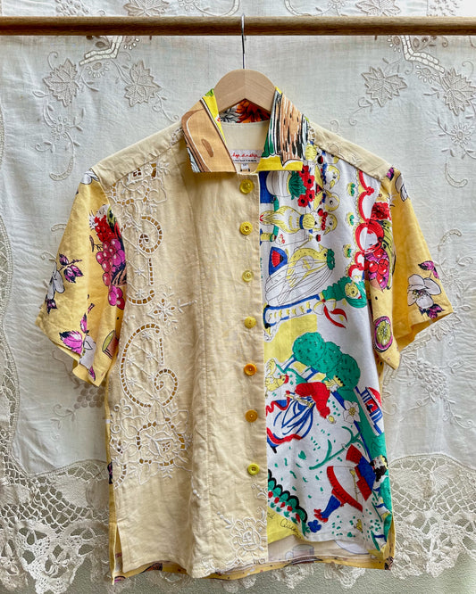 A short-sleeve button-up shirt hanging on a wooden hanger against a white lace curtain backdrop. The shirt features a split design: one side is light beige with embroidered floral motifs, and the other side is white with a vibrant multicolor graphic print.