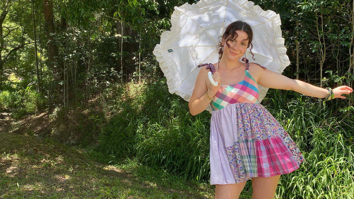 A woman stands outdoors in a wooded area wearing a colorful patchwork mini dress with pastel stripes and plaid patterns, holding a large white flower-shaped umbrella. She is wearing light blue rain boots.