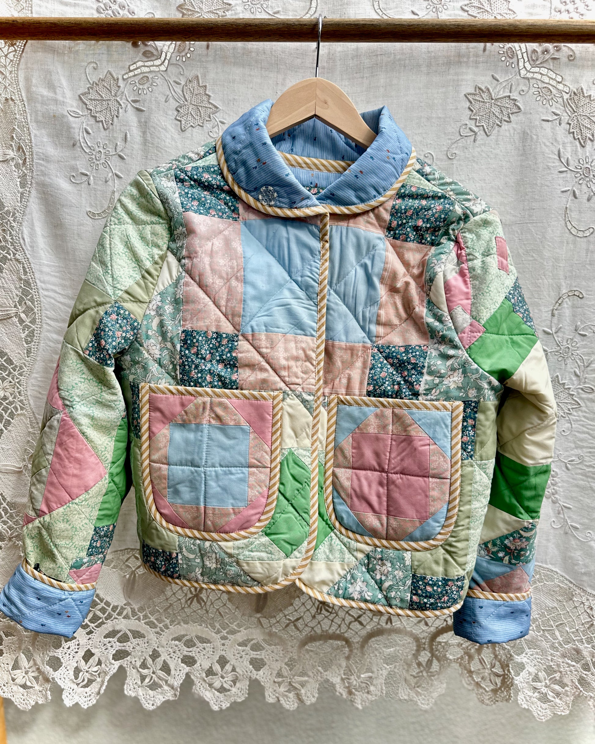 Front view of a multicolored quilted  patchwork jacket made from genuine vintage materials on a hanger against a lace curtain background