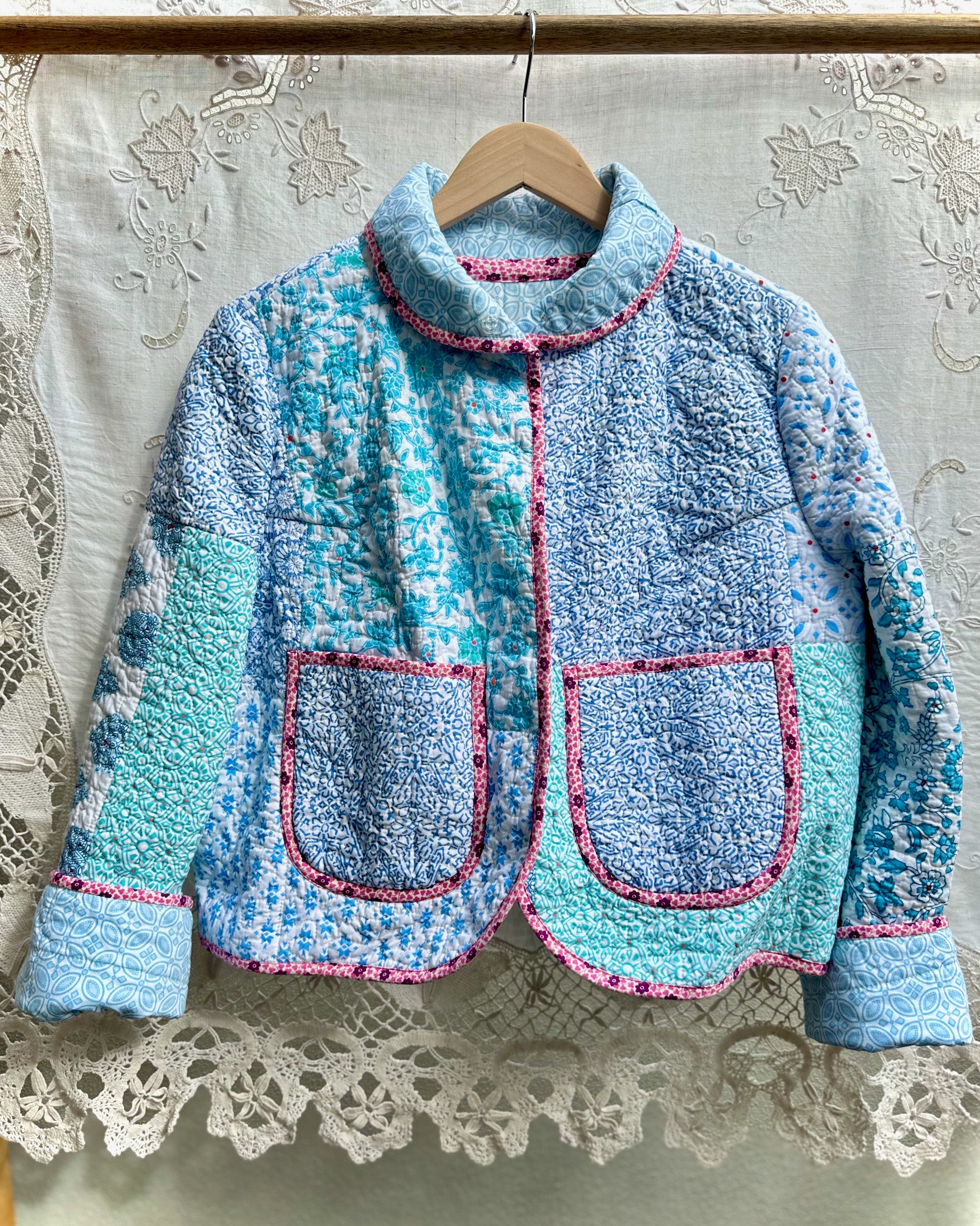 Blue patchwork style jacket with pink liberty of london trim on a hanger against a lace curtain background