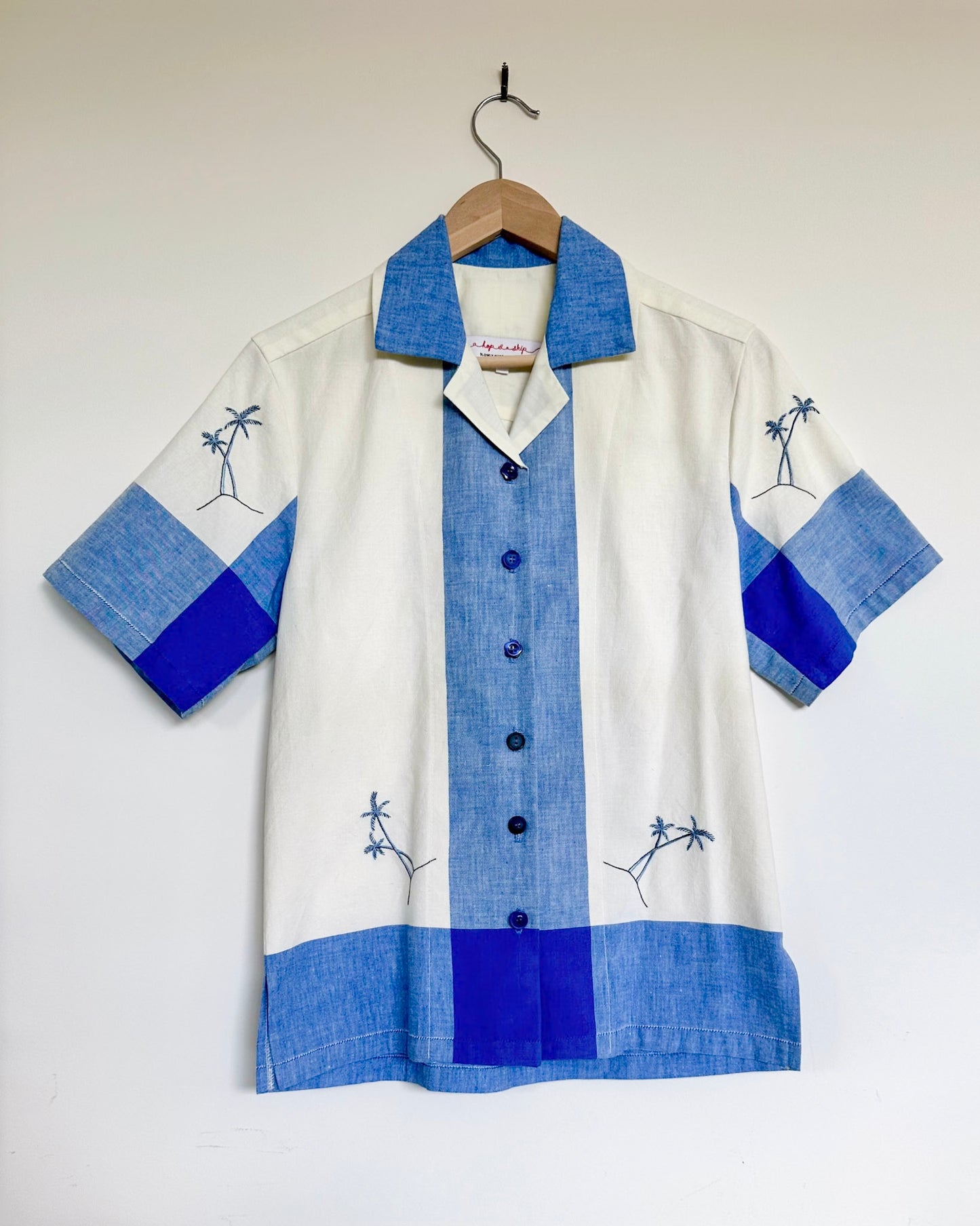 White shirt with blue accents and palm tree designs on a hanger against a white background