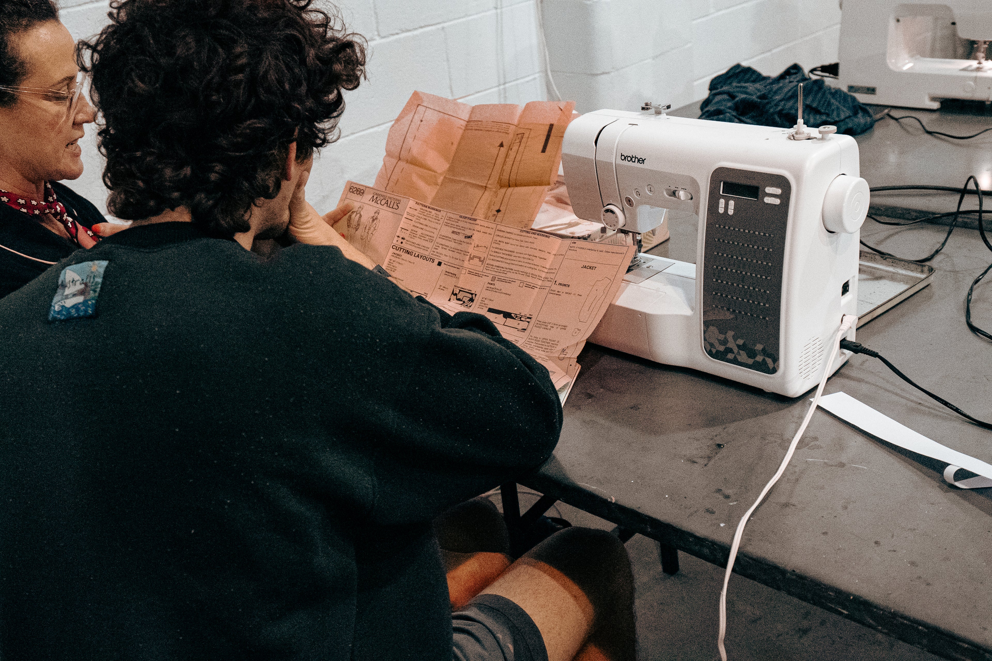 Two people working on a sewing machine with a Brother brand name, surrounded by various sewing items on a table.