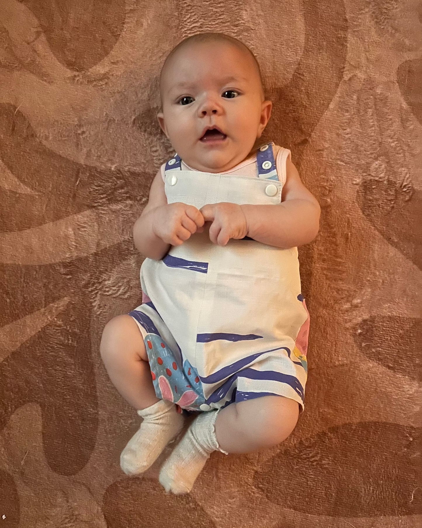 Baby laying on a textured brown surface wearing a white romper with blue accents.